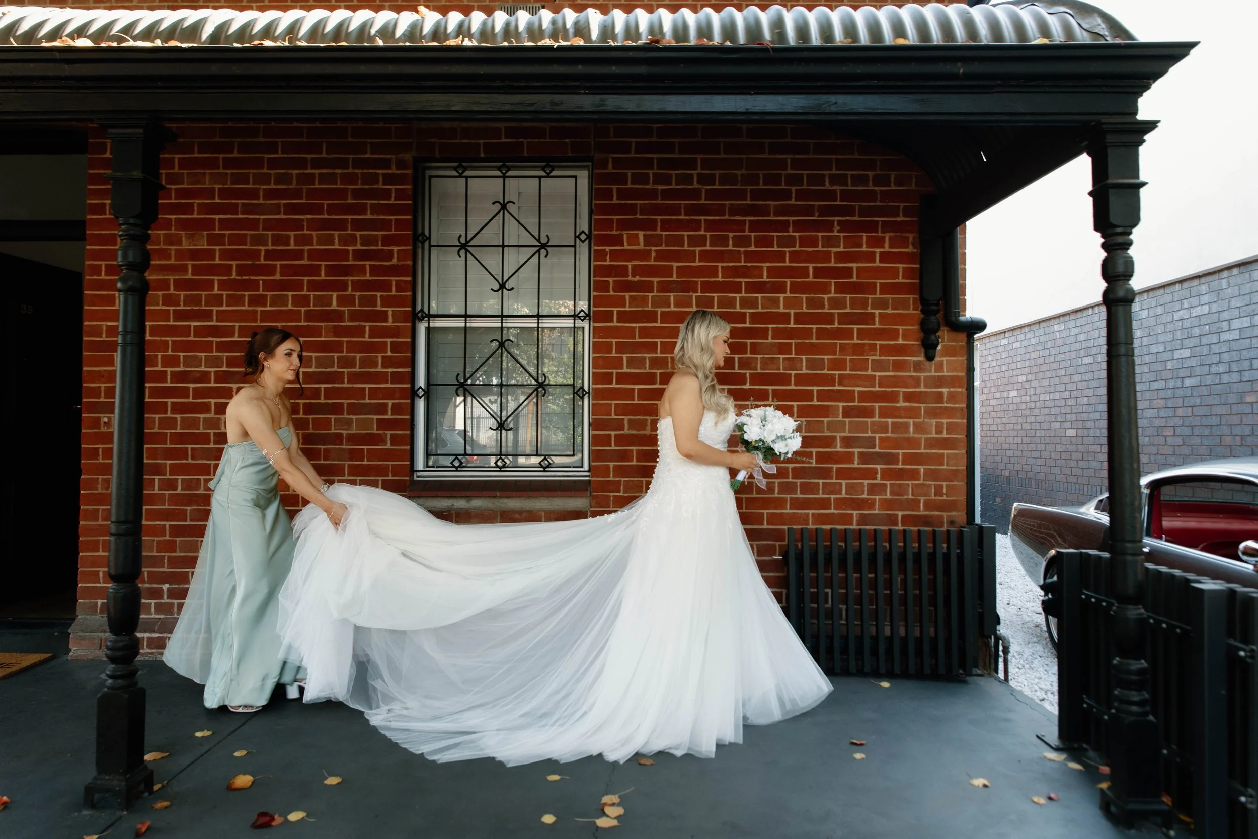 A bride in a white wedding gown holding a bouquet of white flowers, standing on a porch next to a woman in a light blue dress, with a brick wall and window behind them.