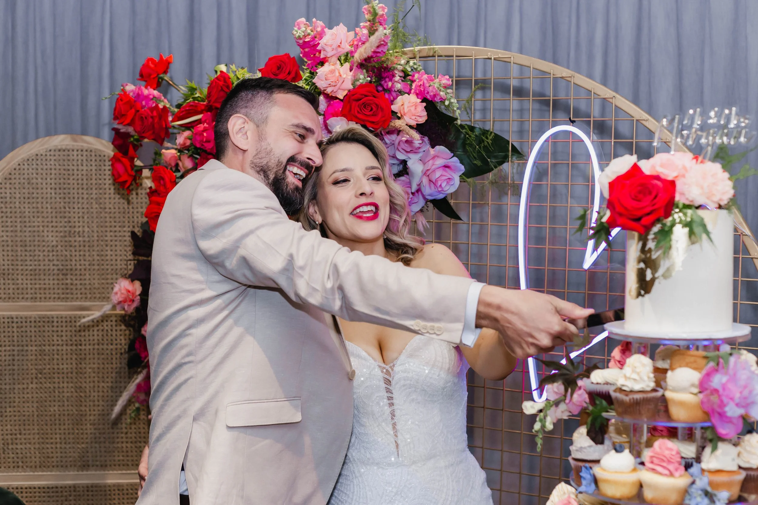 A smiling couple, dressed in wedding attire, cut a wedding cake together at a celebration with floral decorations and a neon heart sign in the background.