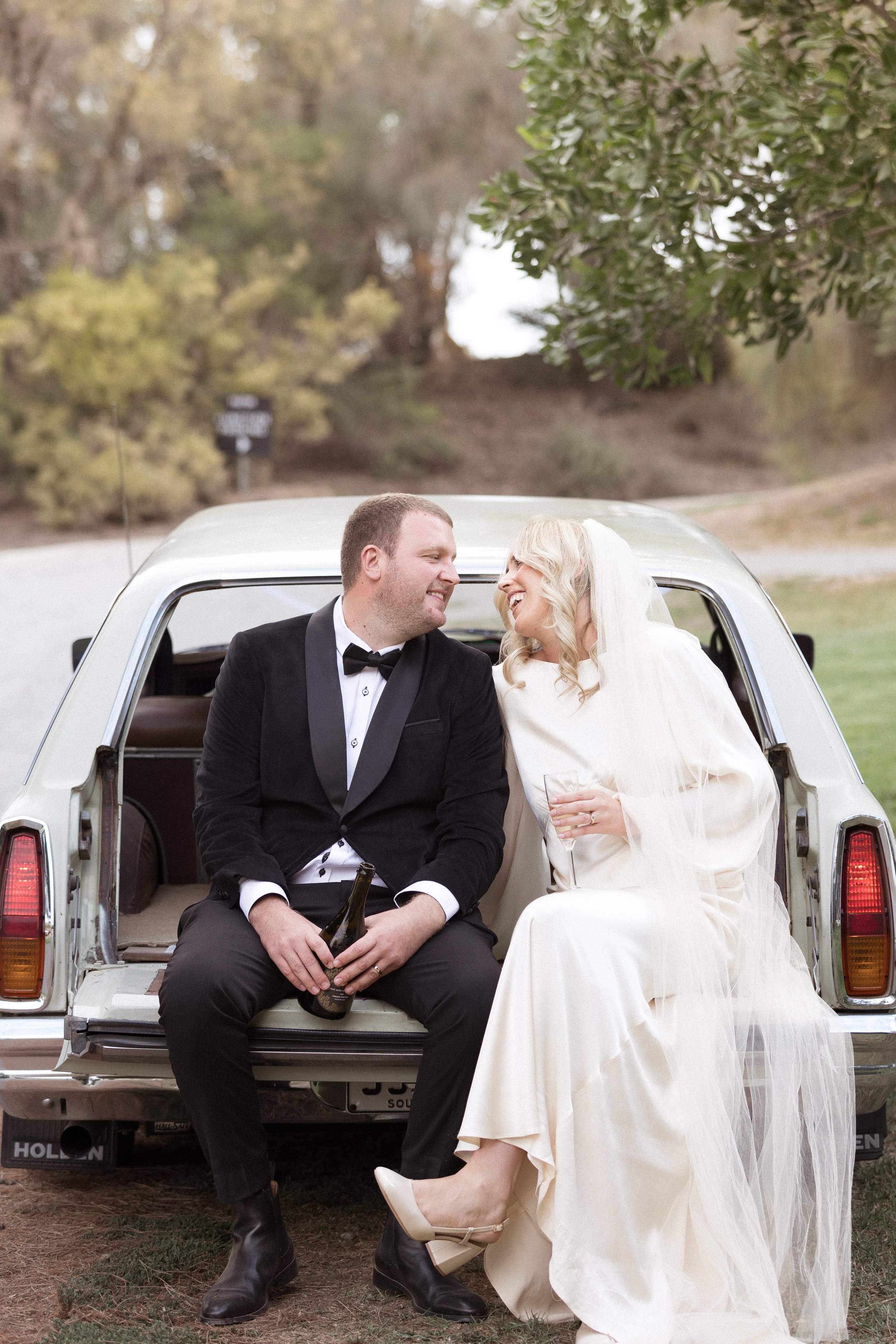 A newlywed couple happily sits in the open trunk of a vintage car, sharing a joyful moment. The groom wears a black tuxedo and holds a beer bottle, while the bride, in a cream-colored wedding dress and veil, holds a glass and smiles at him. They are 