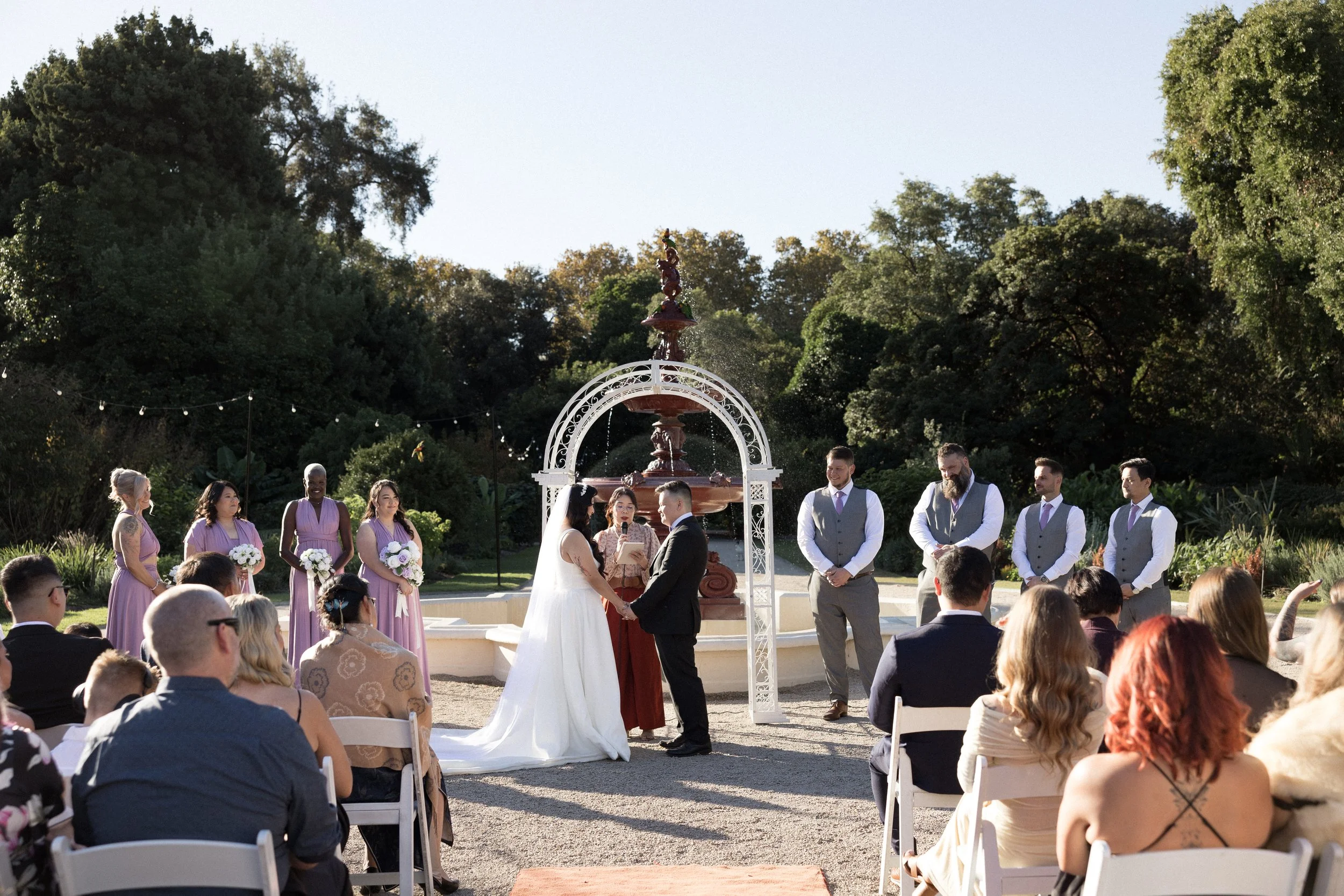 Bride and groom exchanging vows at an outdoor wedding ceremony in front of a decorative fountain and an arch, with bridesmaids and groomsmen standing nearby, surrounded by guests seated on white chairs in a lush garden setting.