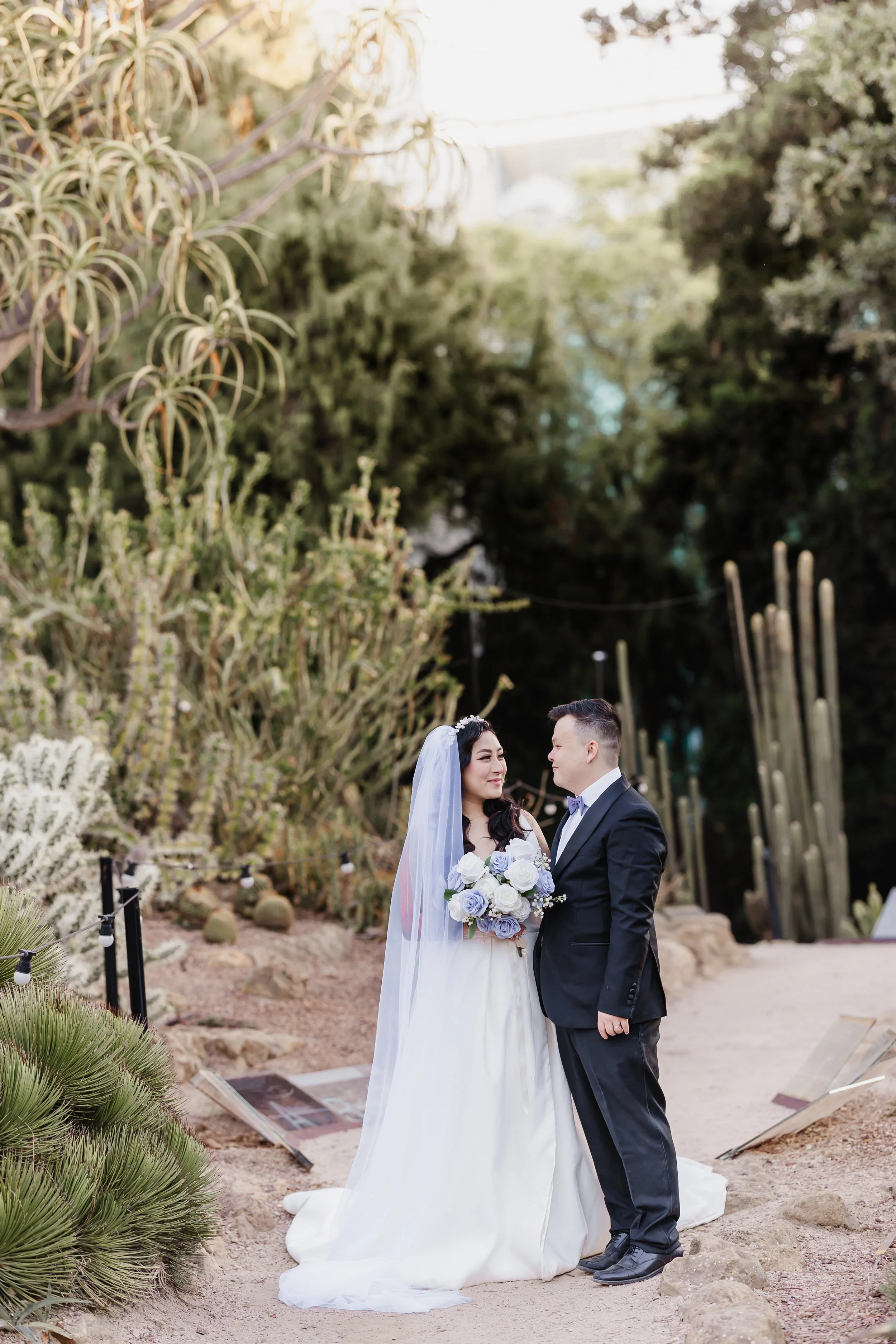 A bride and groom standing together outdoors on their wedding day, surrounded by desert plants and cacti.