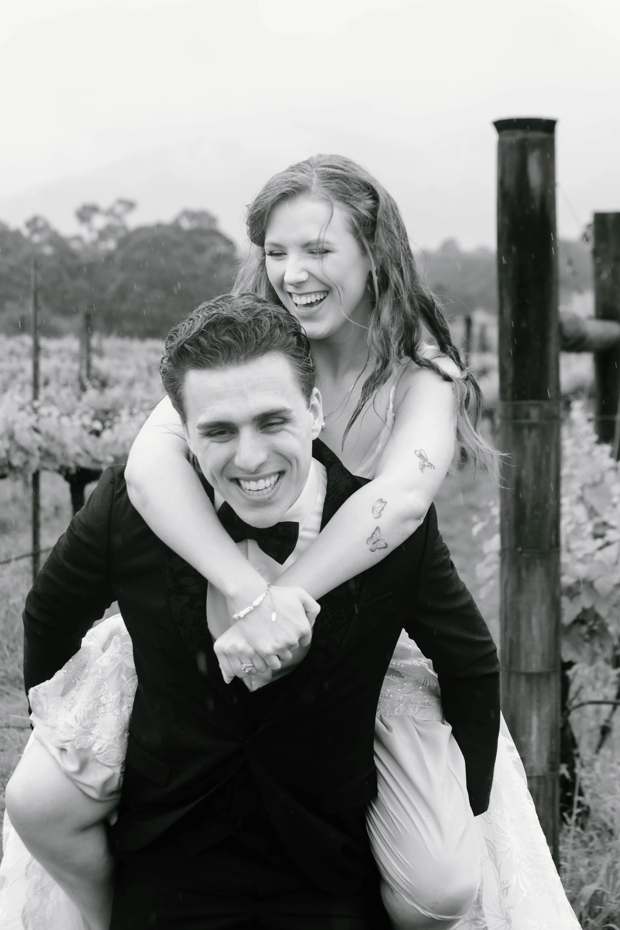 A black and white photo of a joyful couple outdoors, with the woman on the man's back, both smiling and laughing, in a vineyard setting.