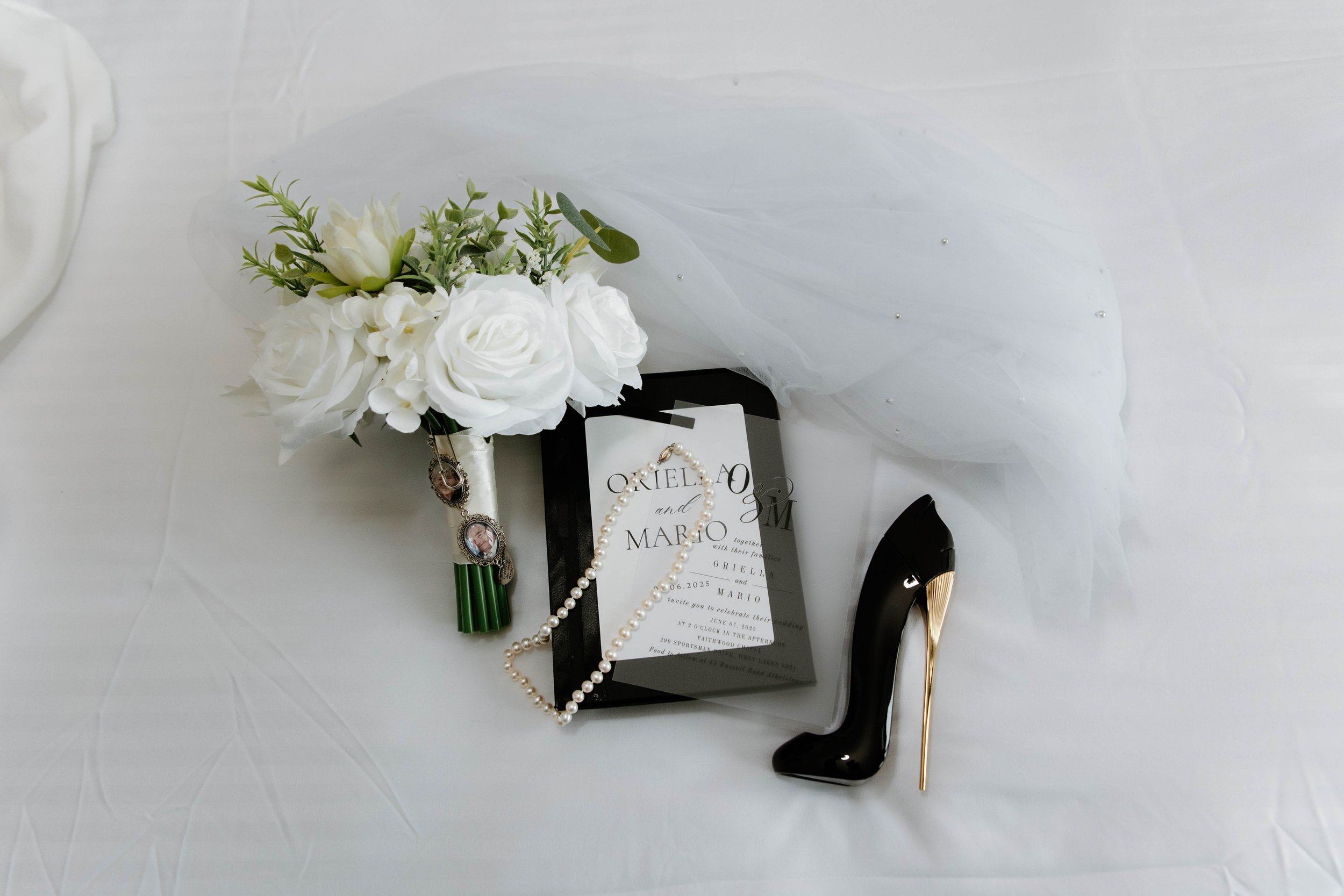 Wedding bouquet with white roses and greenery, a pearl necklace, a black high-heeled shoe with a gold heel, and a wedding invitation with a mirror frame on a white fabric surface.
