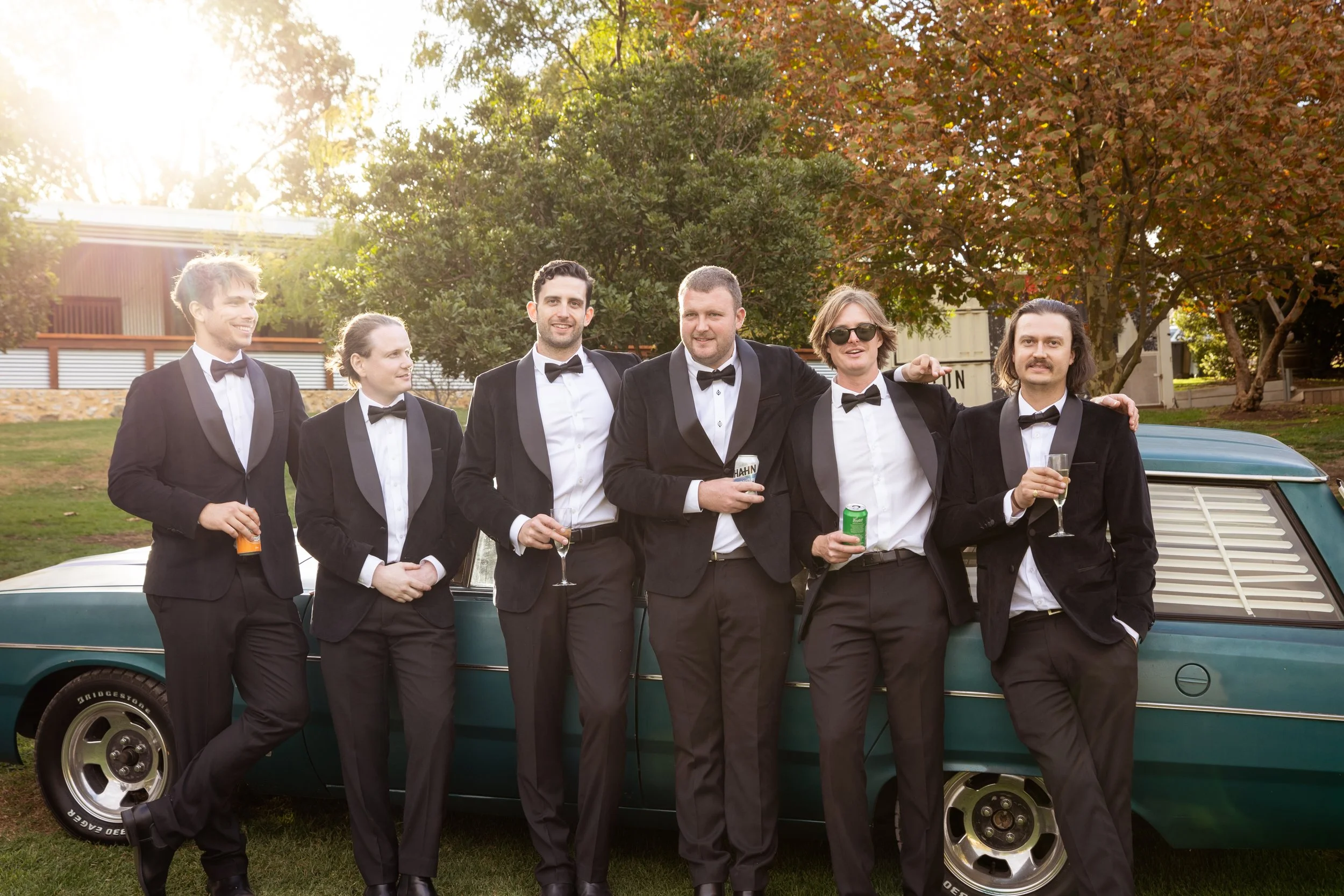 Group of six men in tuxedos with bow ties and drinks, standing in front of a vintage car outdoors during sunset.
