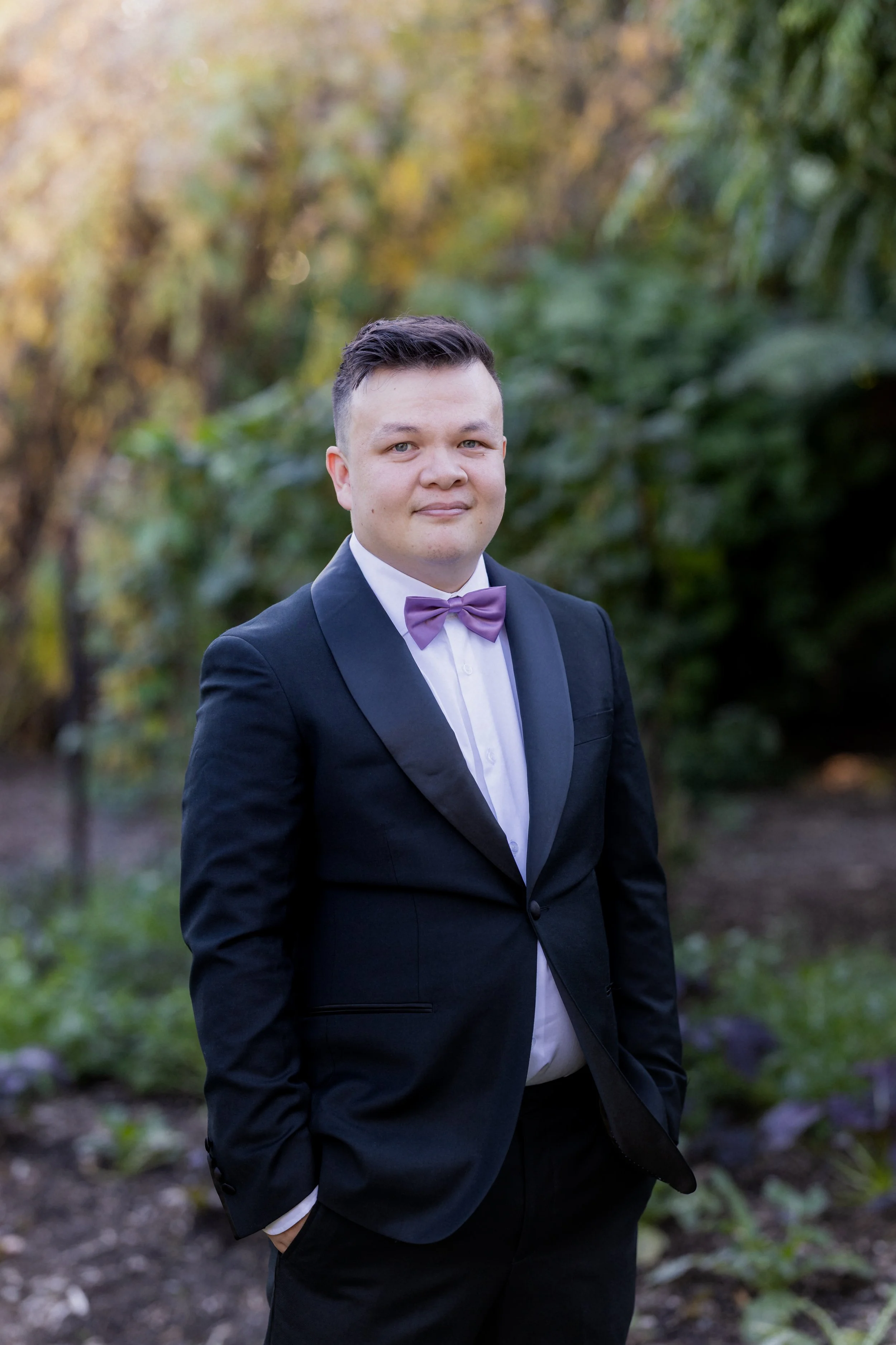A man in a black tuxedo with a purple bow tie standing outdoors in a garden with trees and plants in the background.