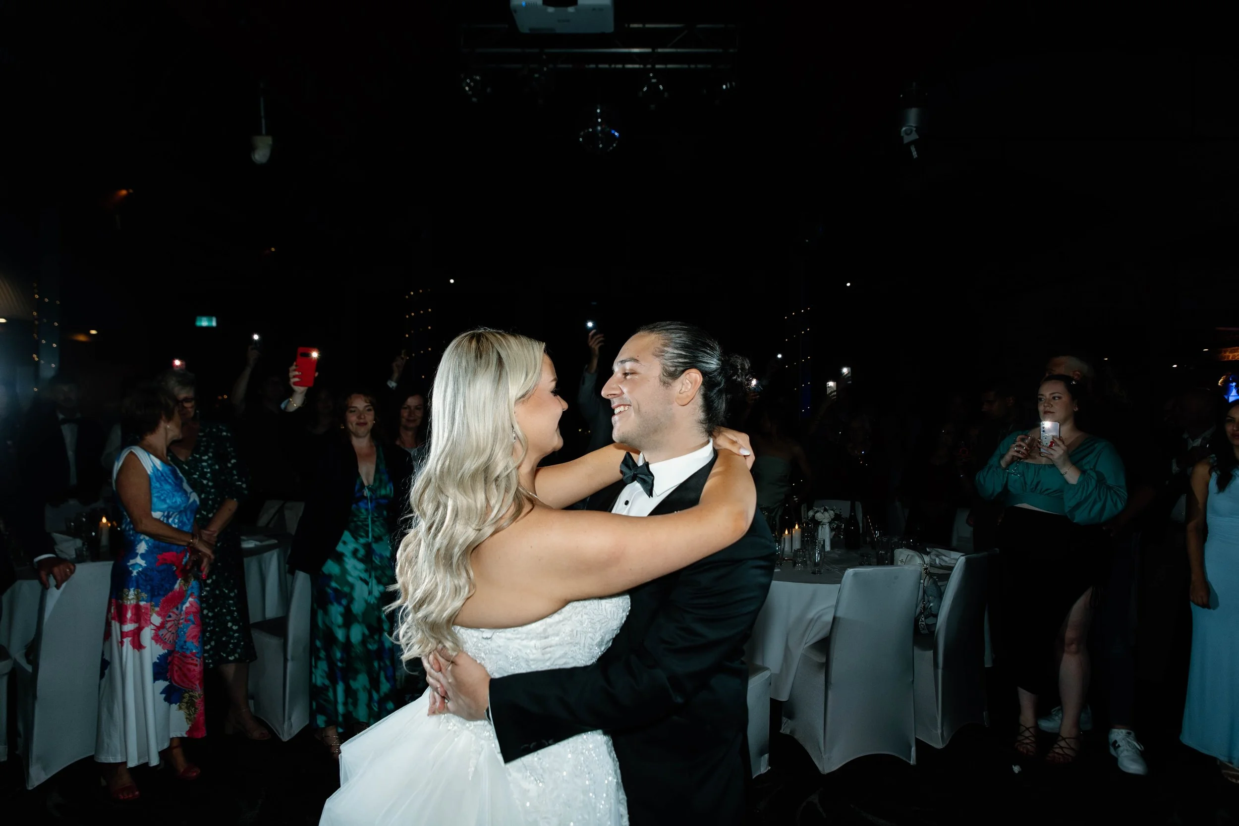 A bride and groom dance at their wedding reception surrounded by guests, some taking photos, in a dimly lit venue.