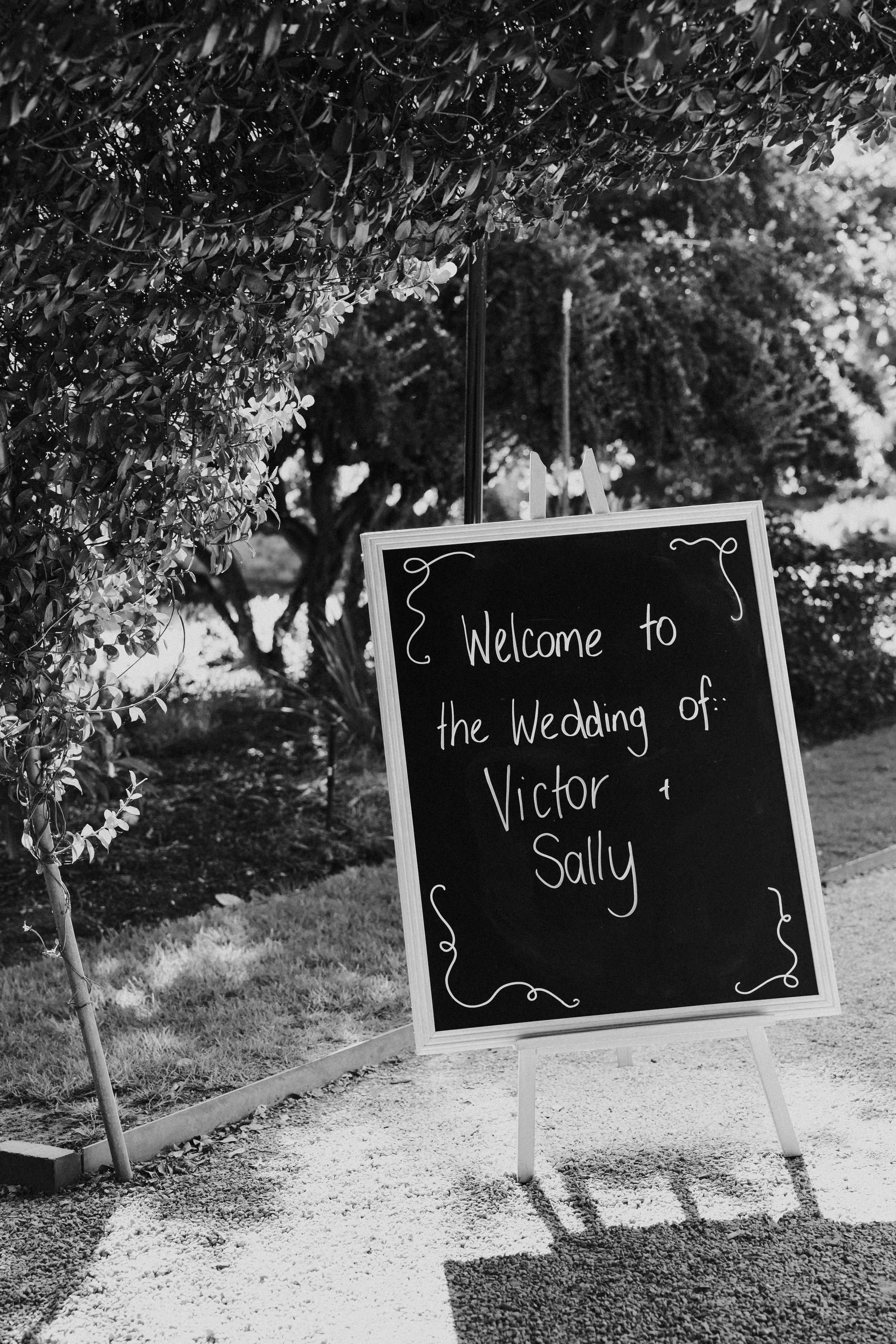 A blackboard sign at an outdoor wedding reception welcomes guests to the wedding of Victor and Sally, surrounded by trees and greenery, with a shadow cast on the gravel path.