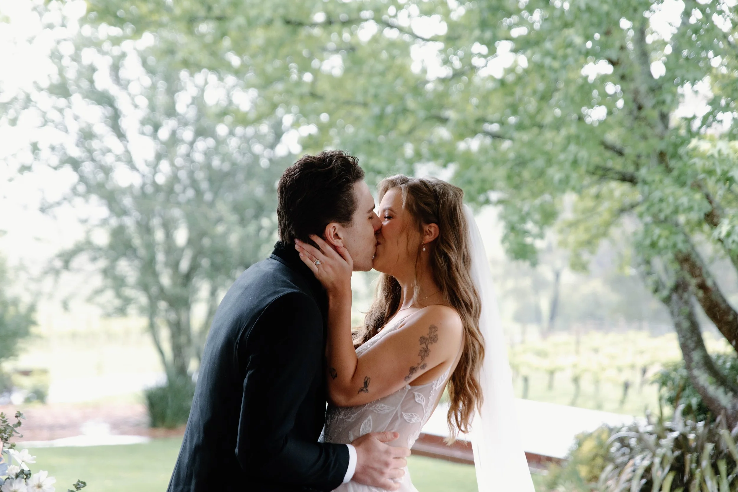 A bride and groom sharing a kiss outdoors during their wedding ceremony, with lush trees in the background.