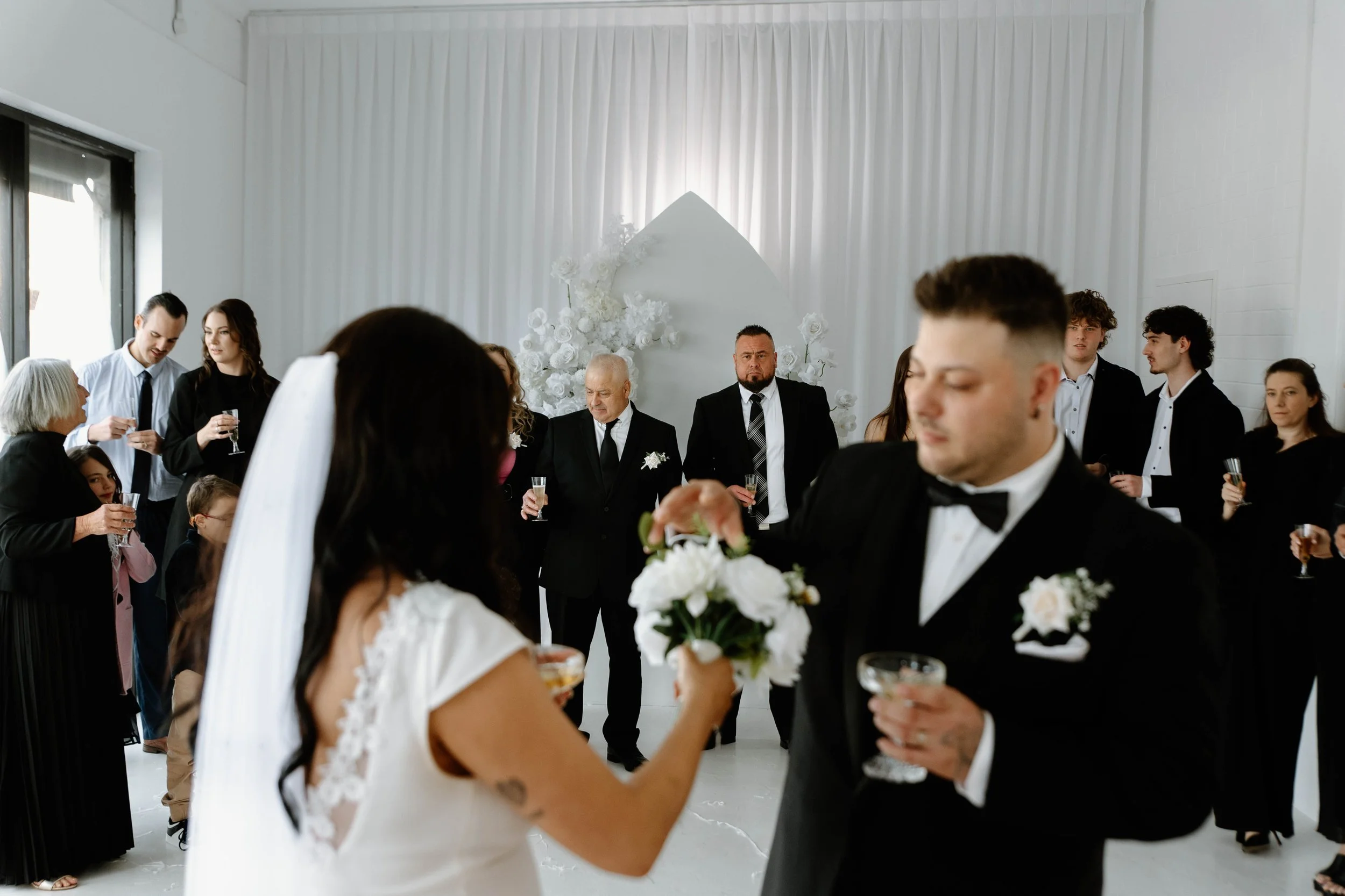 A wedding reception with the bride and groom in the foreground, the bride holding a bouquet and the groom holding a drink, surrounded by guests dressed in formal attire, in a white decorated room.