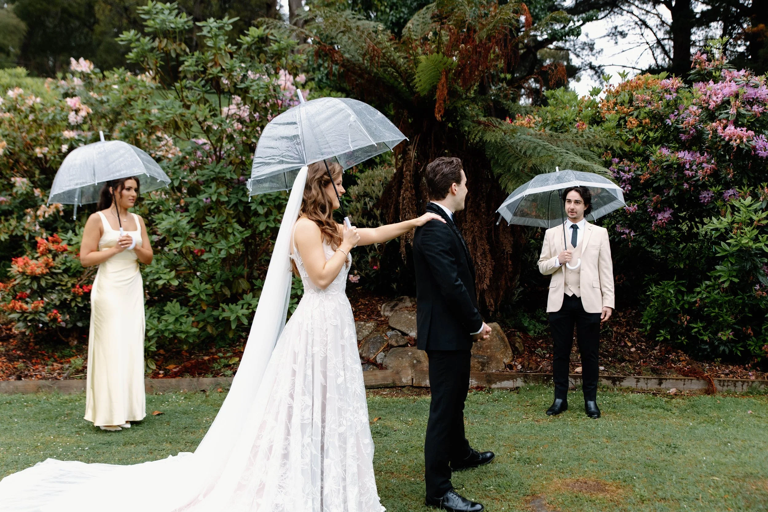 A wedding ceremony outdoors in the rain with four people holding transparent umbrellas, including a bride in a white gown, a groom in a black suit, a bridesmaid in a cream dress, and a man in a beige jacket.