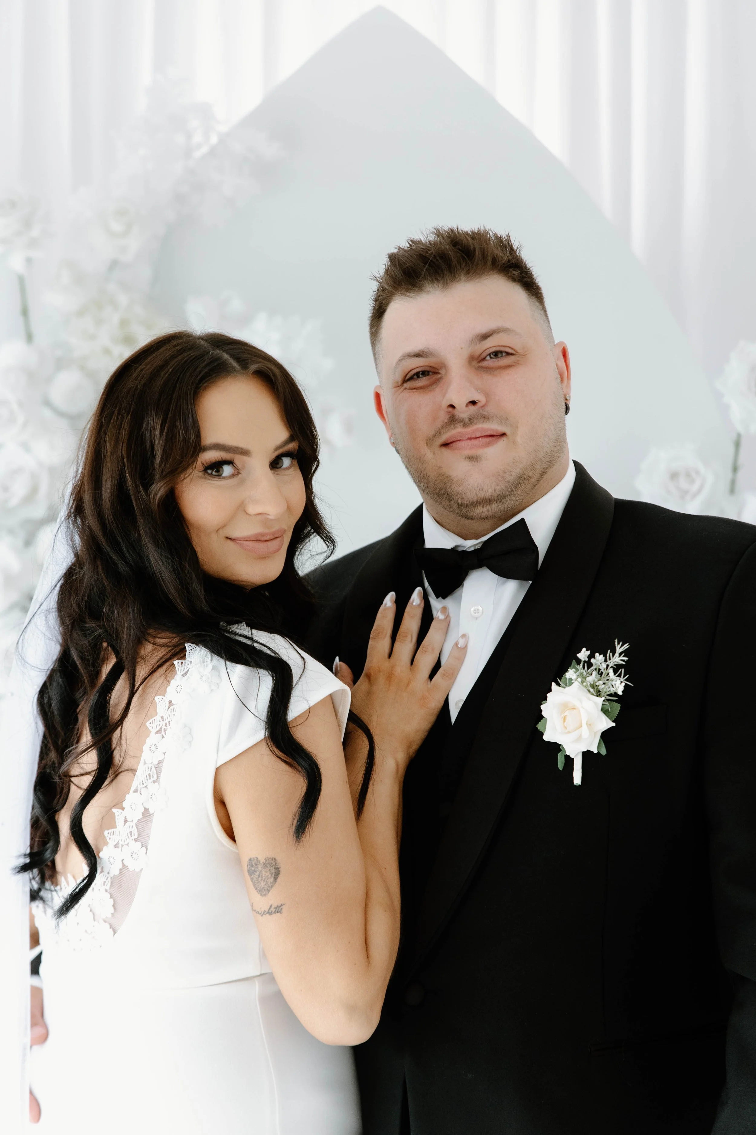 A bride and groom posing for a wedding photo indoors. The bride has long dark hair, a white dress with lace details, and a tattoo on her arm. The groom is dressed in a black tuxedo with a bow tie and a white boutonniere. They are standing close toget