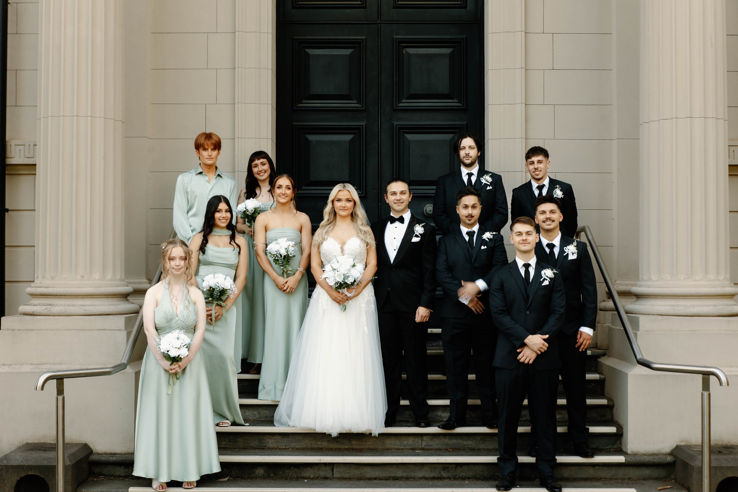 A group of people standing on the steps of a building, dressed in formal attire for a wedding, with women in green dresses and men in black tuxedos, some holding bouquets of flowers.