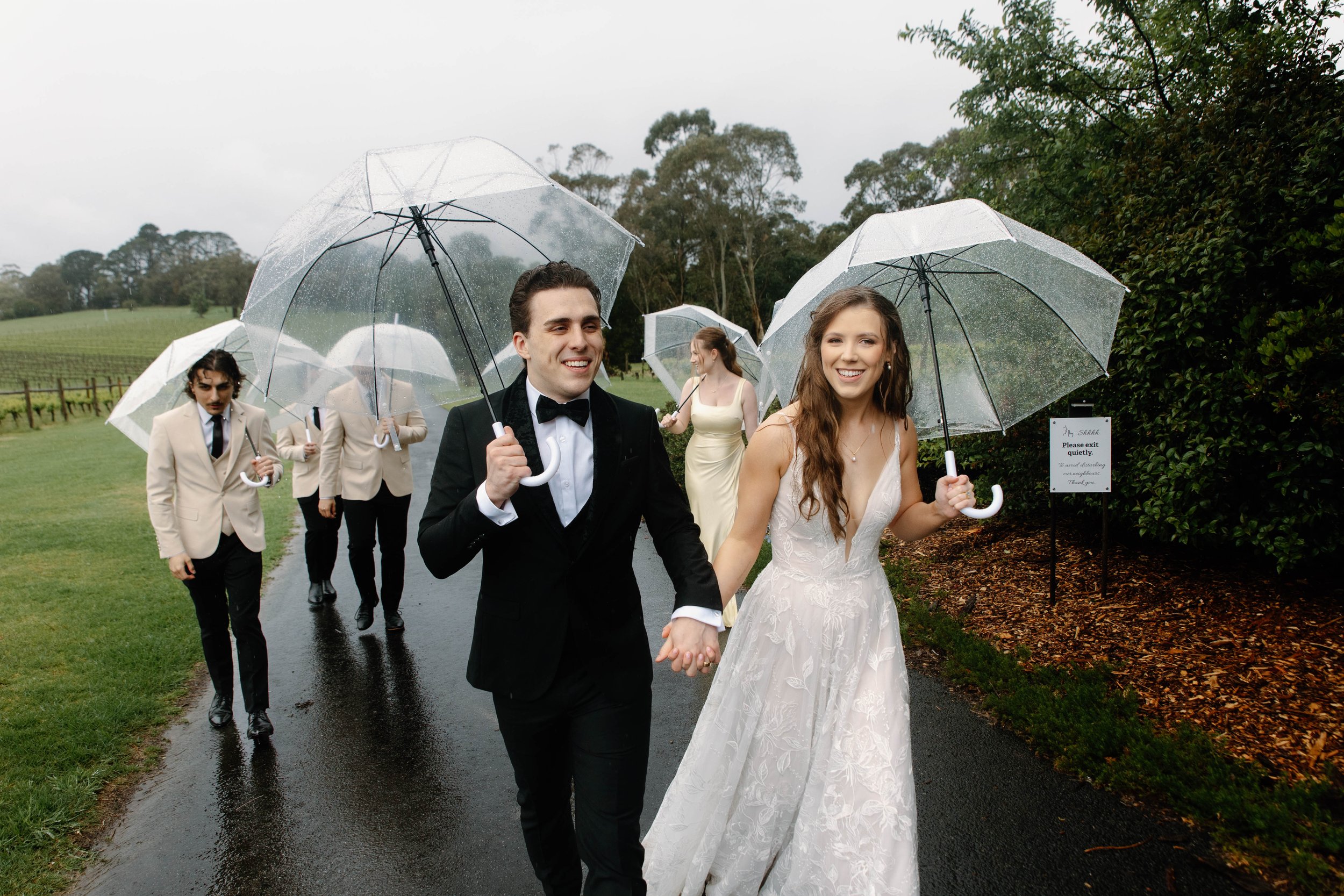 Wedding couple holding umbrellas and walking on a rainy day outdoors, with guests behind them, trees, and a vineyard in the background.