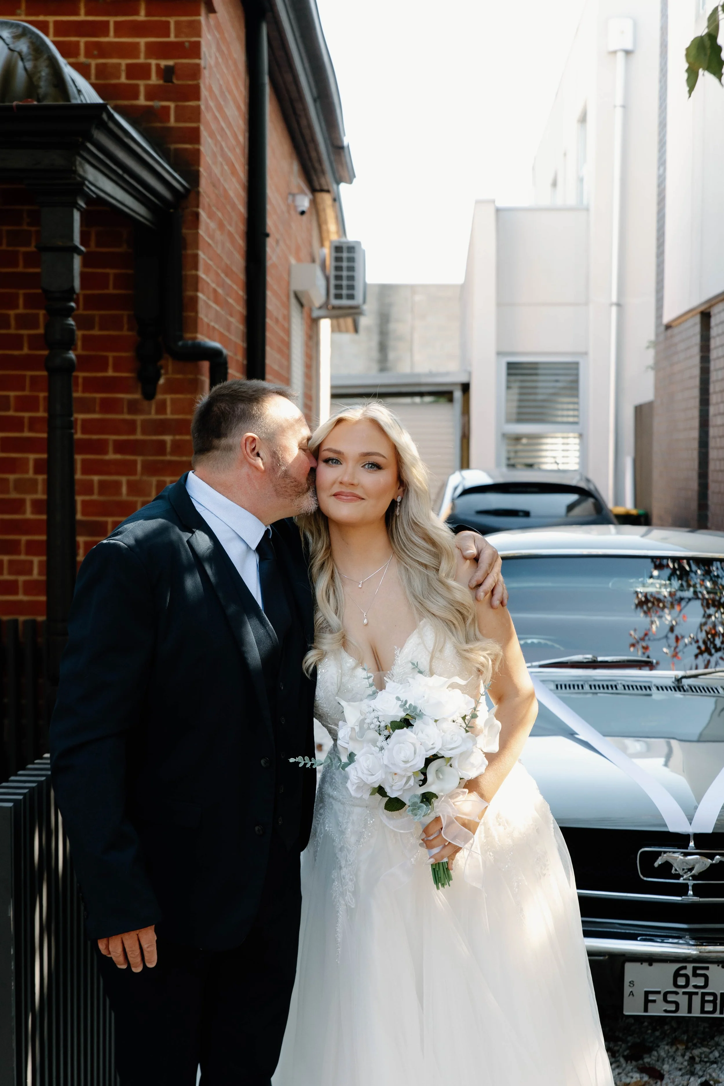 A bride holding a bouquet of white roses and lilies, standing next to a man in a dark suit who is kissing her cheek, in an outdoor setting with cars and buildings in the background.
