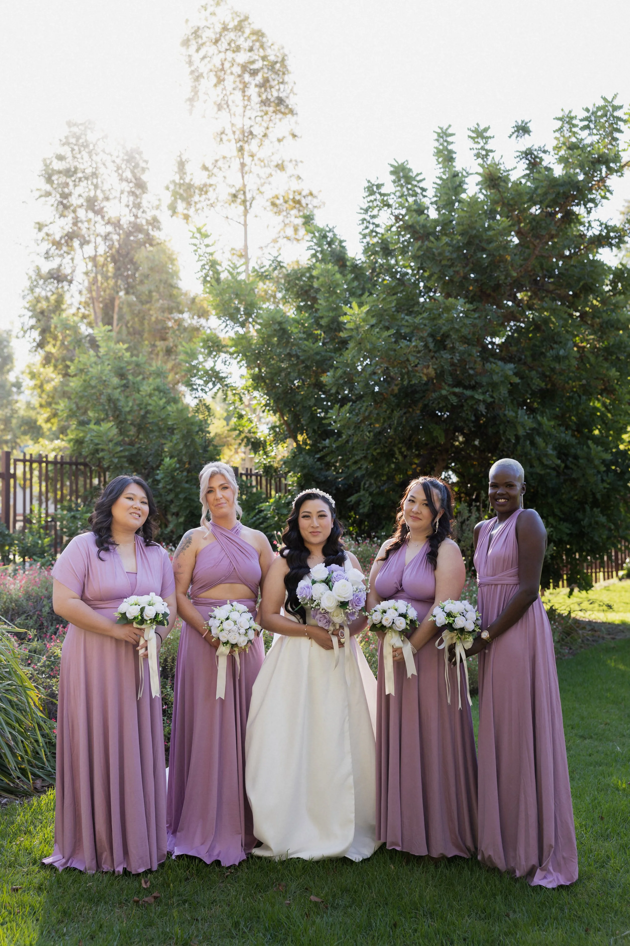 A bride and four bridesmaids standing outdoors in a garden, holding bouquets of white and purple flowers, with green trees and sunlight in the background.