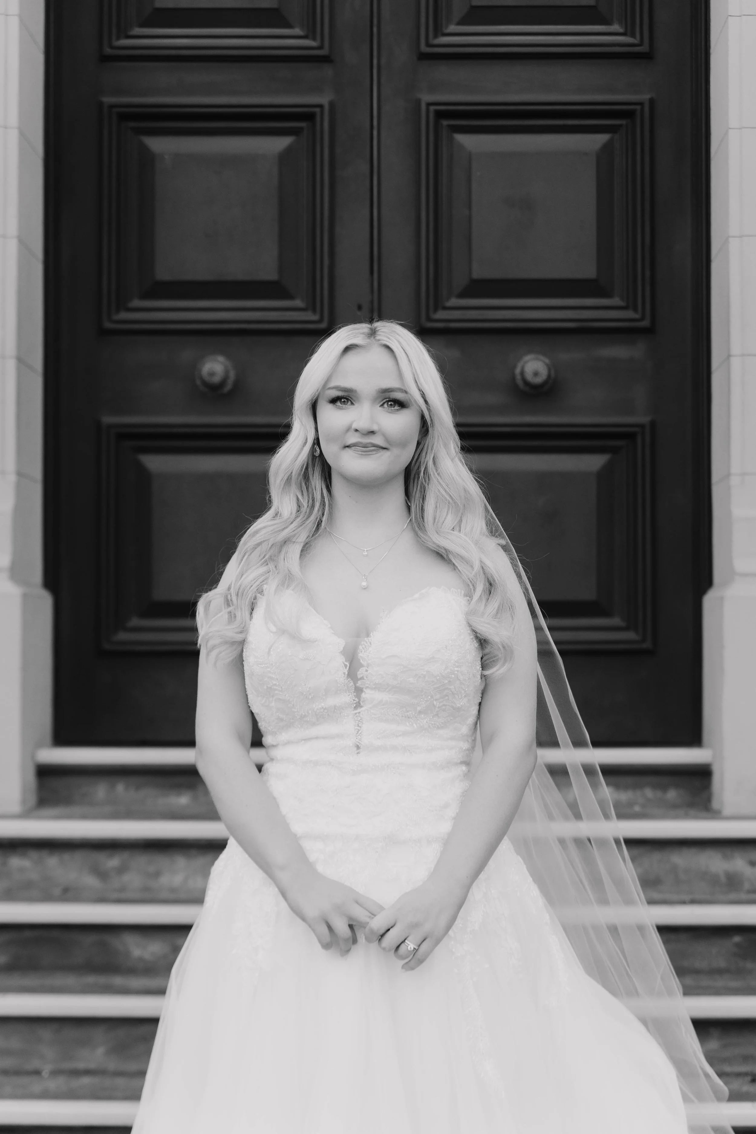 A woman in a wedding dress standing on the steps of a building with large dark wooden doors.
