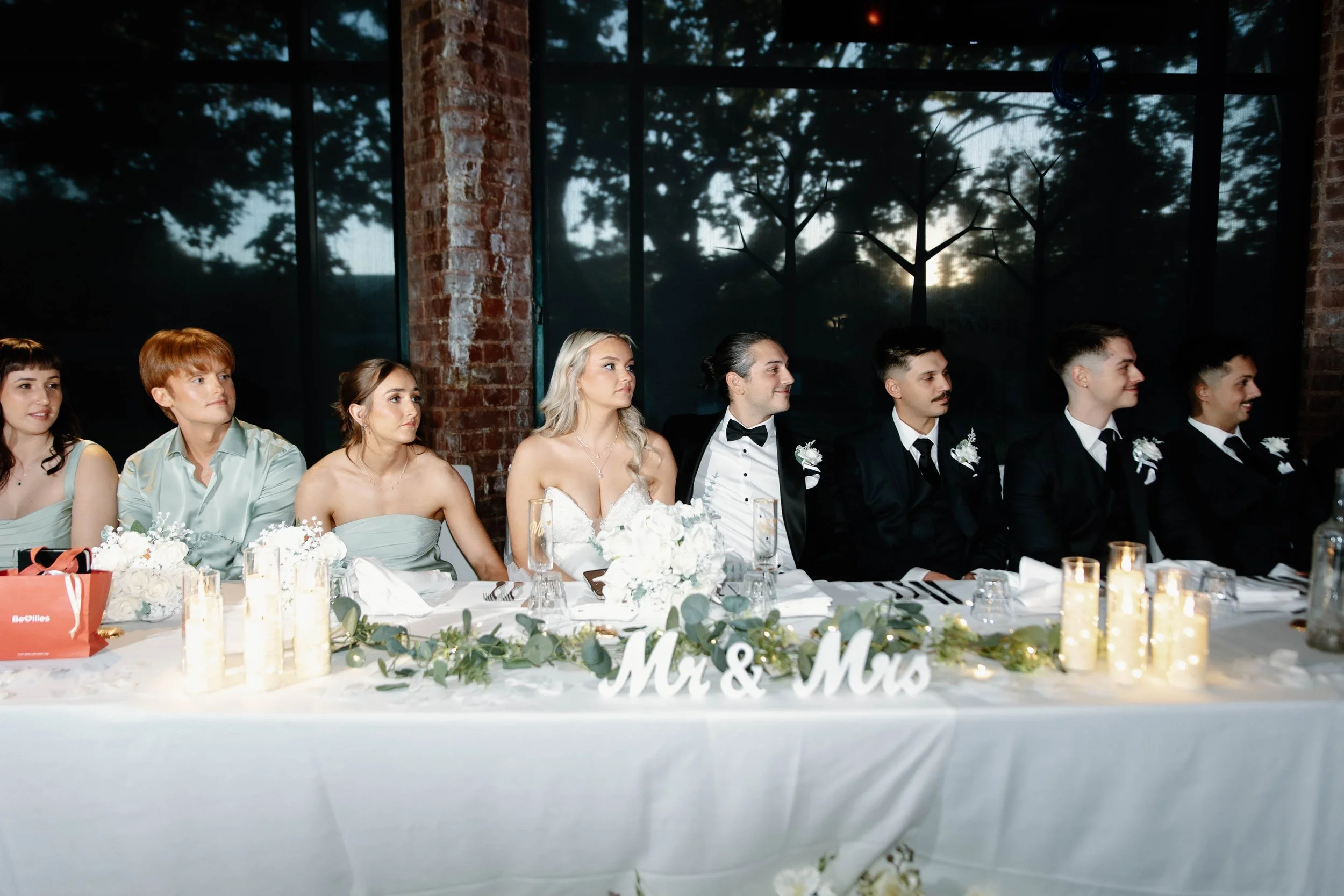 A group of people sitting at a wedding reception table, dressed in formal attire, with a rustic brick wall and large window in the background.