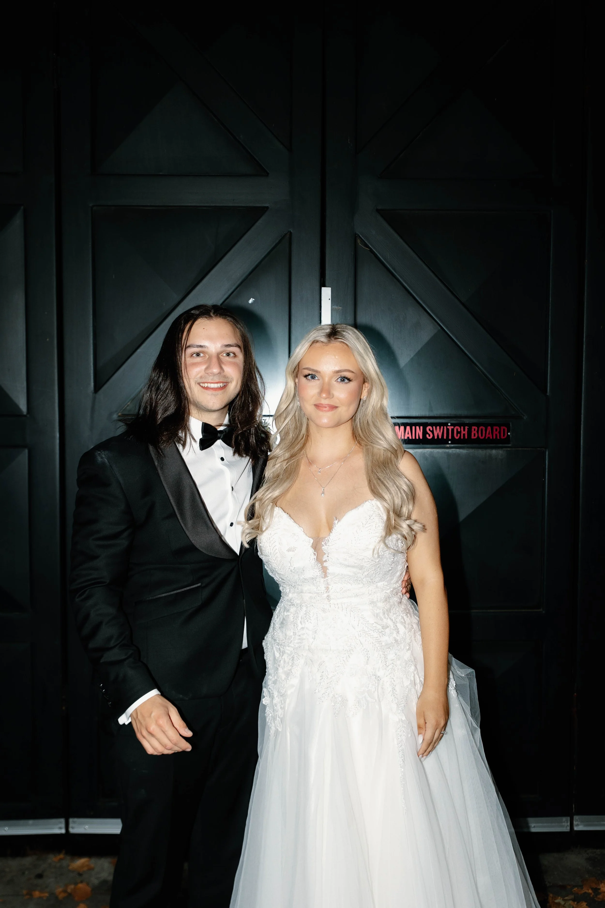 A man in a tuxedo and a woman in a white wedding dress standing together in front of large black doors with red lettering that says 'Main Switch Board'.