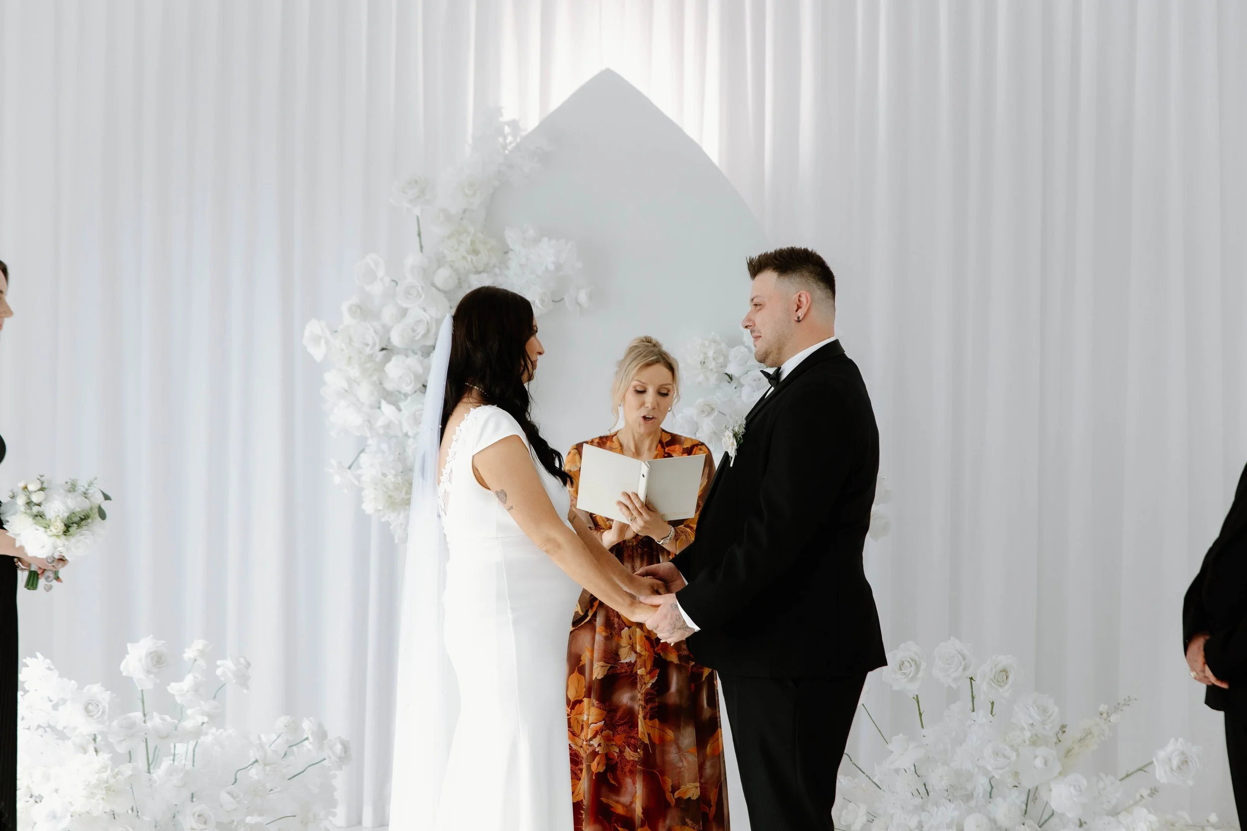 A couple getting married in a white ceremony, holding hands and standing before a woman reading vows, with white floral decorations and a white backdrop.