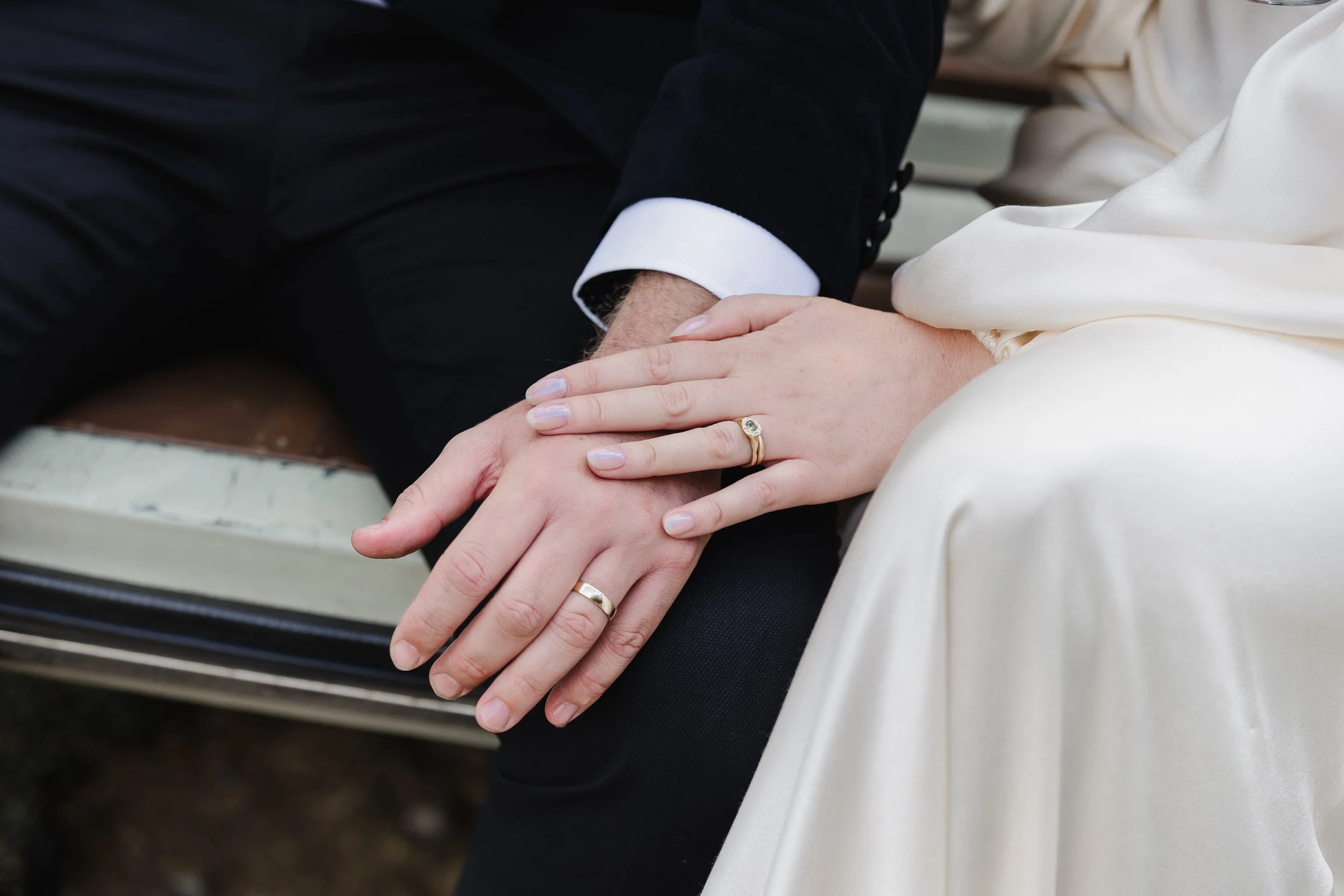 Close-up of a couple's hands with wedding rings resting on a bench, the bride's hand on top of the groom's hand, both wearing wedding bands.