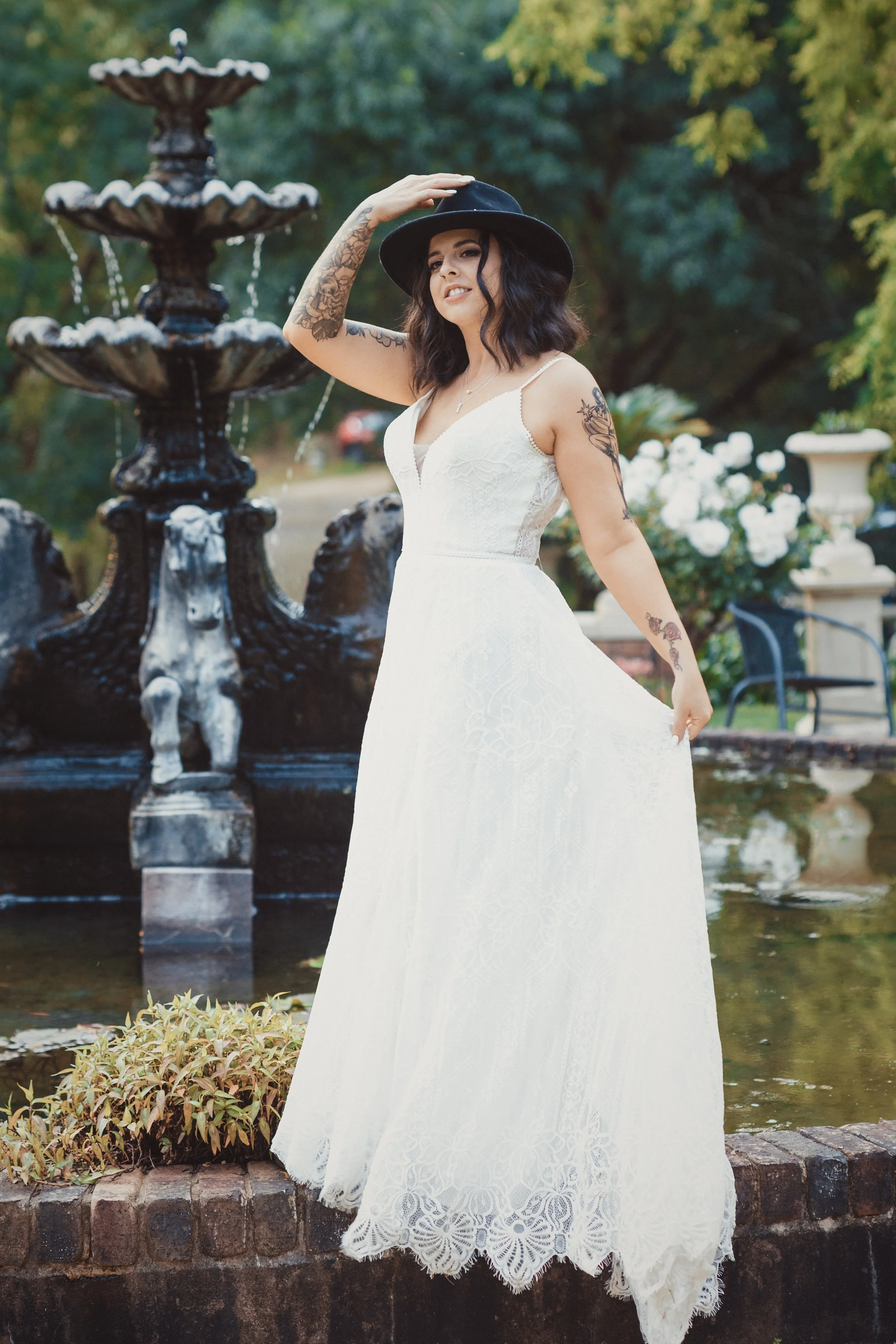 Woman in a white lace dress and black hat standing next to a fountain in a park.