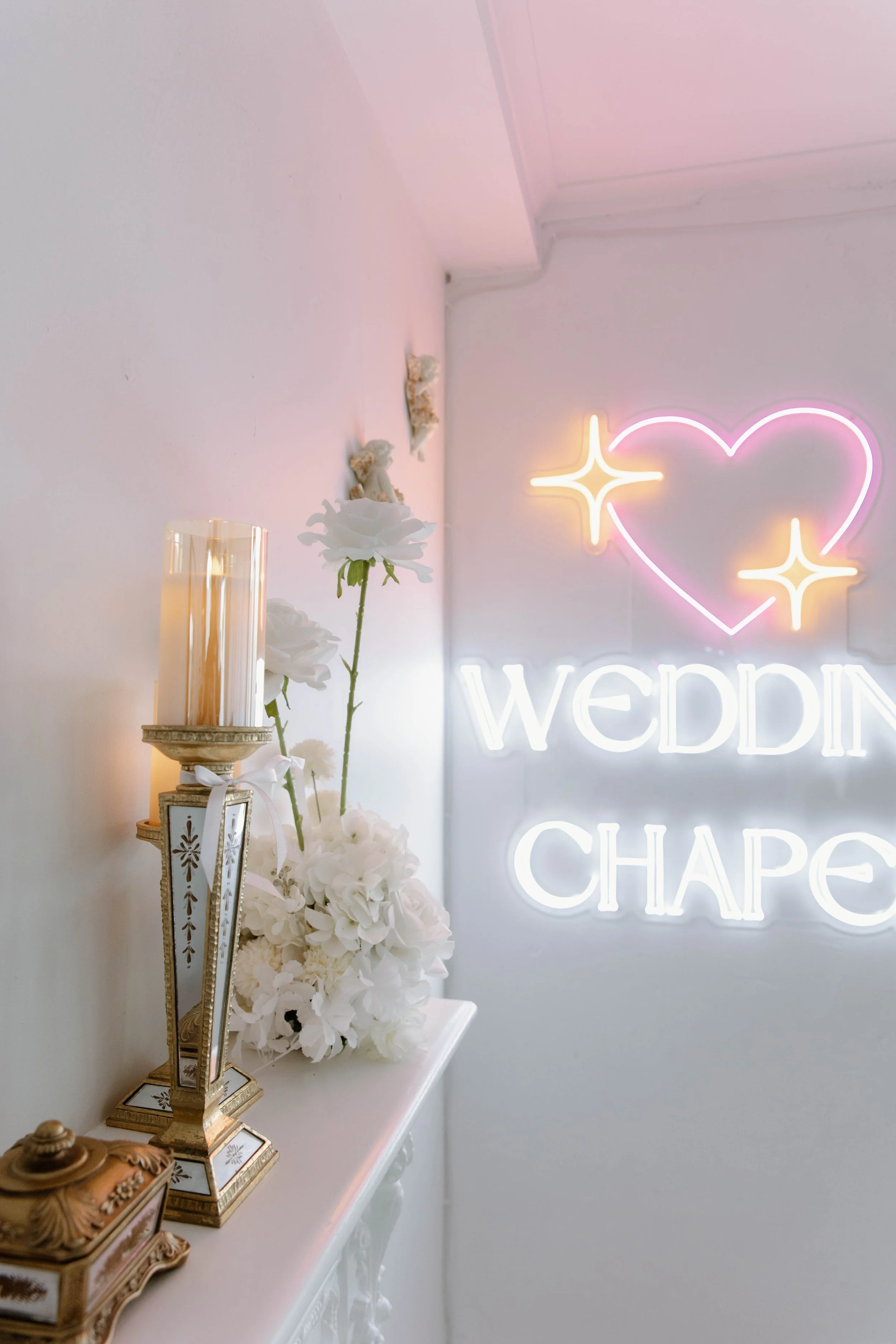 Decorative wedding chapel interior with a white floral arrangement, candles, and a neon sign reading "wedding chapel" with a heart and star accents.