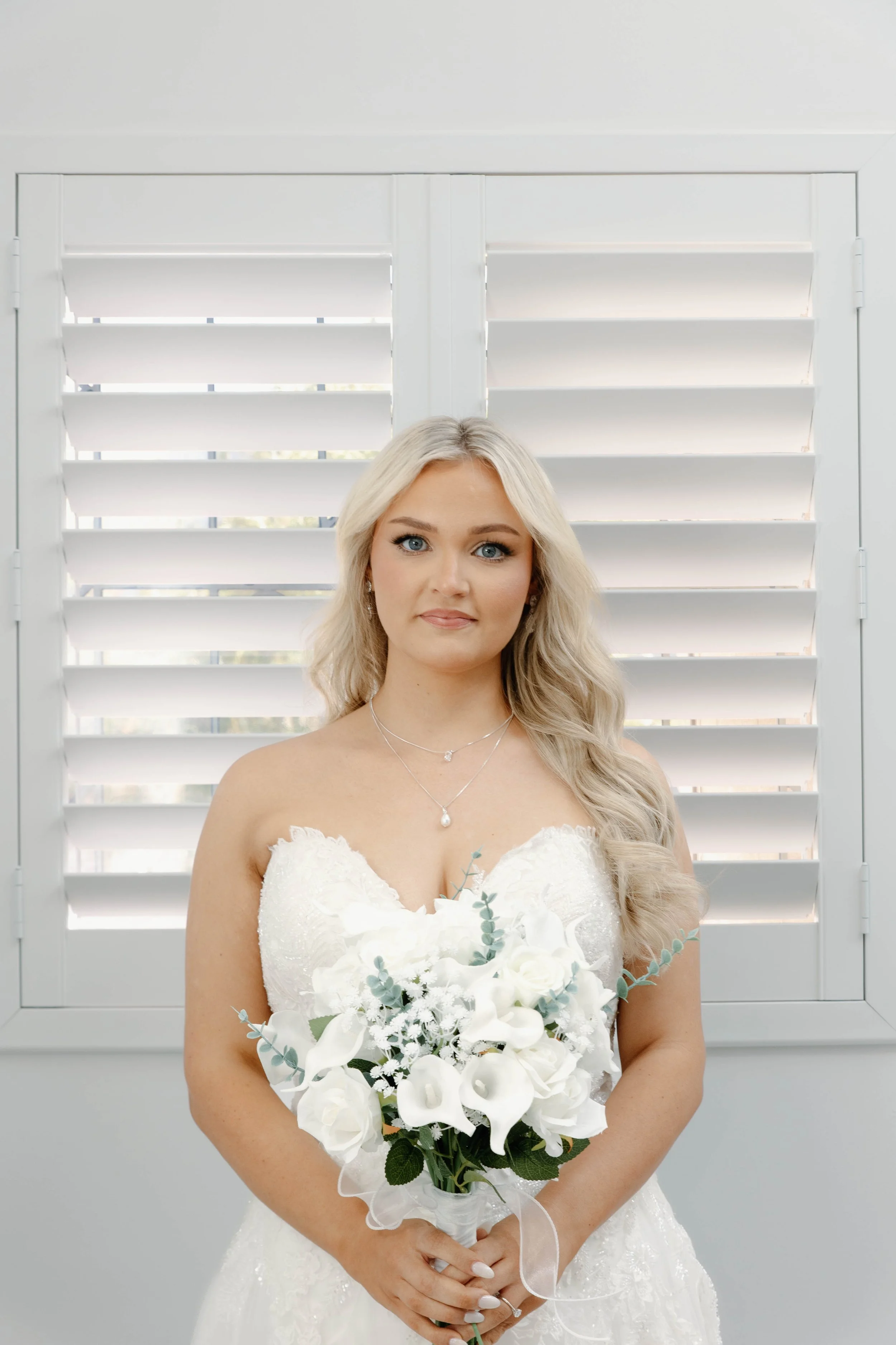 A bride with long blonde hair in a white wedding dress holding a large bouquet of white flowers, standing in front of white plantation shutters.