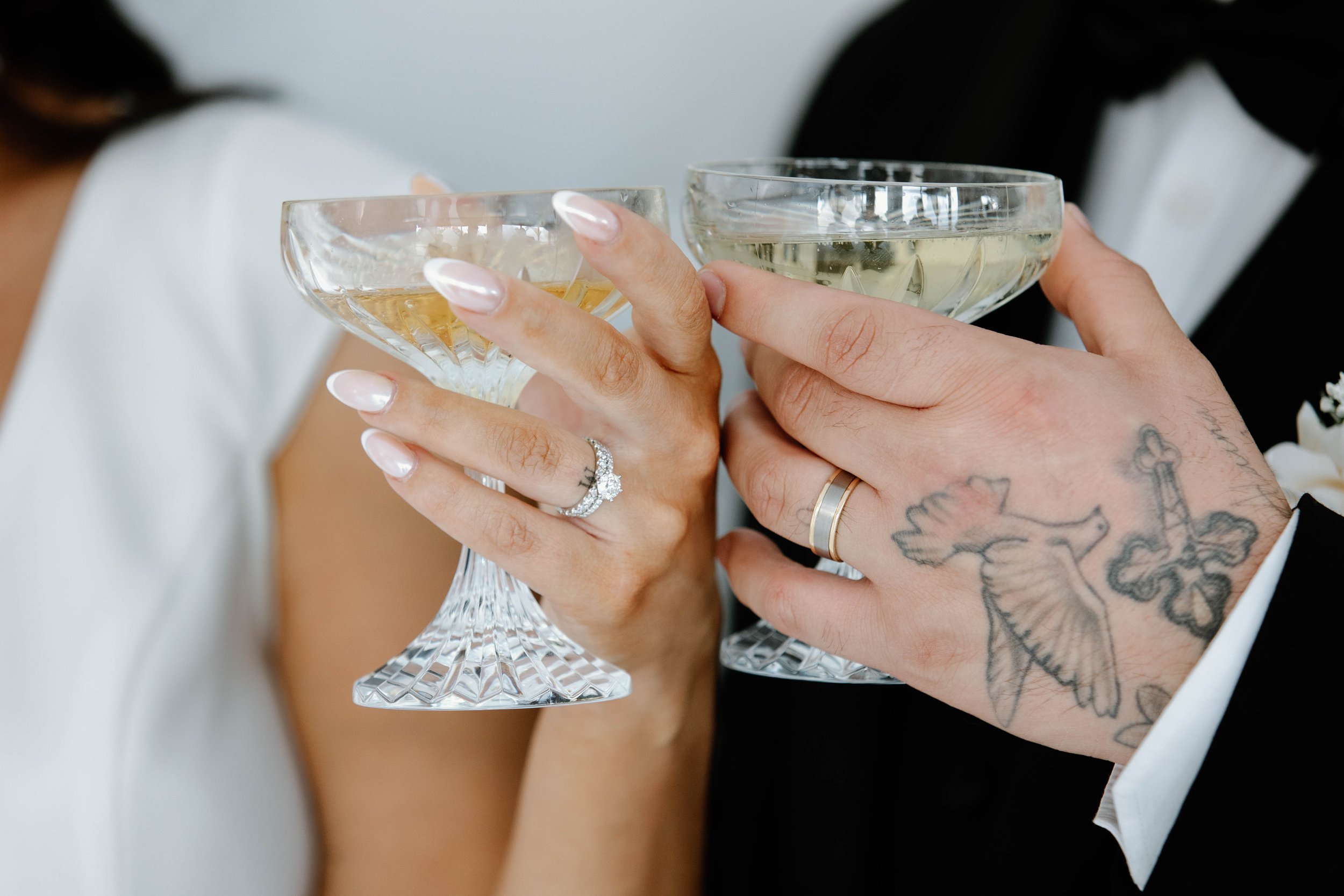 A close-up of a bride and groom toasting with champagne glasses at their wedding, showing the bride's ring and the groom's arm tattoos.