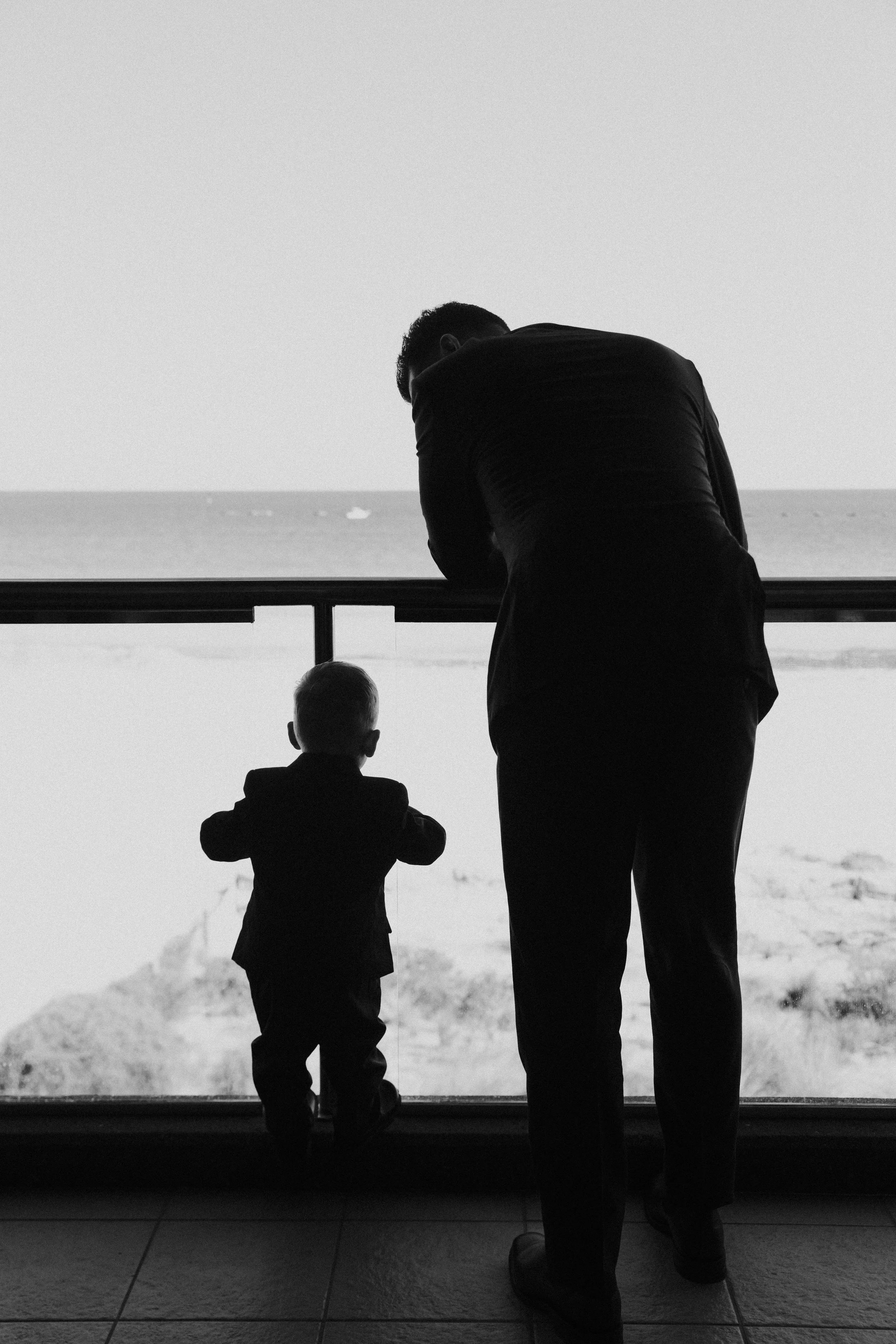 A man and a young boy in suits stand by a window, overlooking the beach and ocean, with the man leaning on the window frame and the boy gazing out.