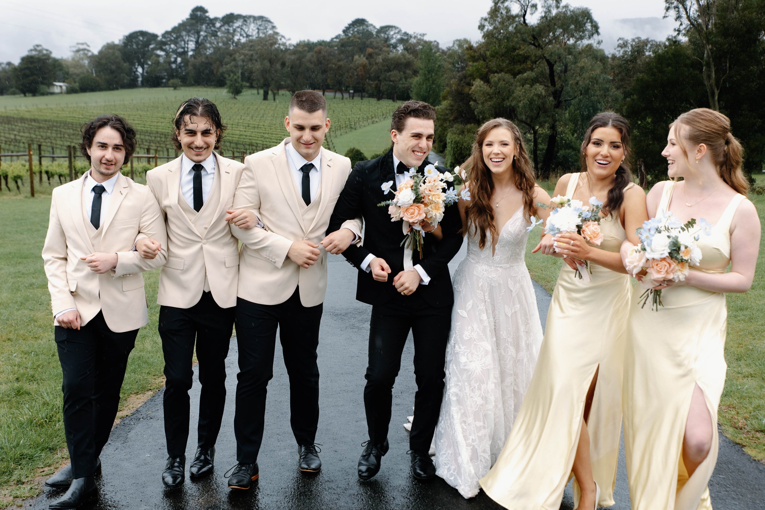 A wedding party walking outdoors on a wet path, with four men dressed in beige suits and black ties, and three women in yellow dresses and a bride in a white gown holding bouquets, all smiling.