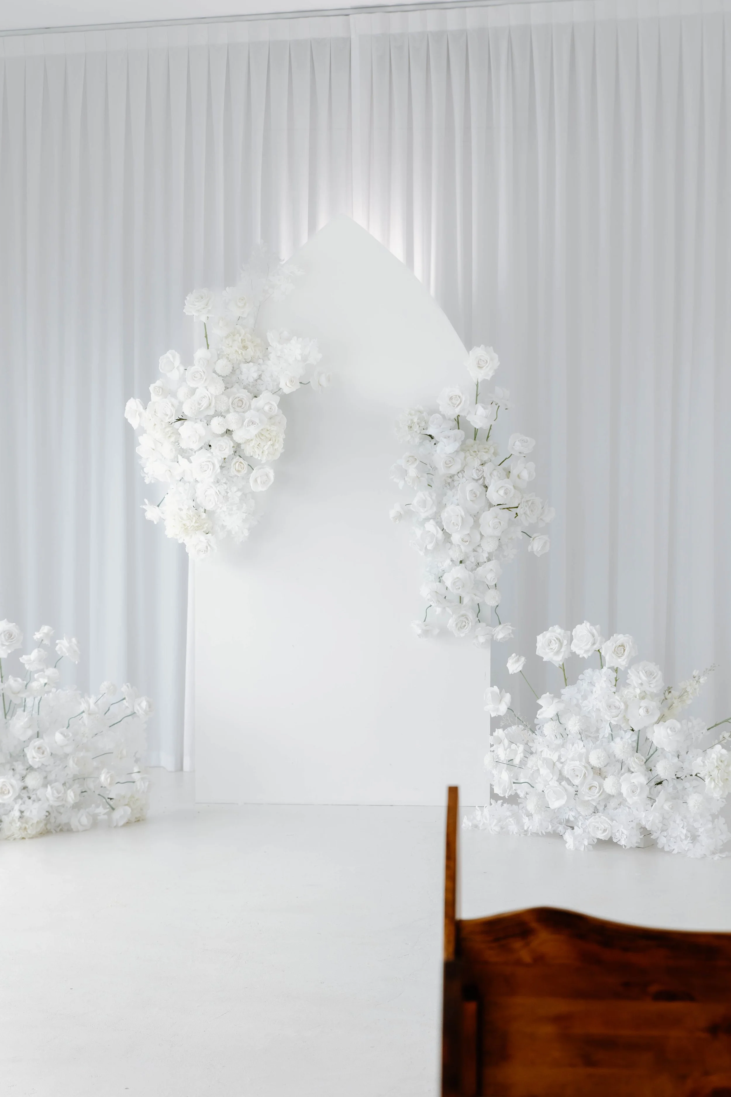 Elegant white wedding ceremony backdrop with floral arrangements of white roses and flowers, white draped curtains, and a wooden bench in the foreground.
