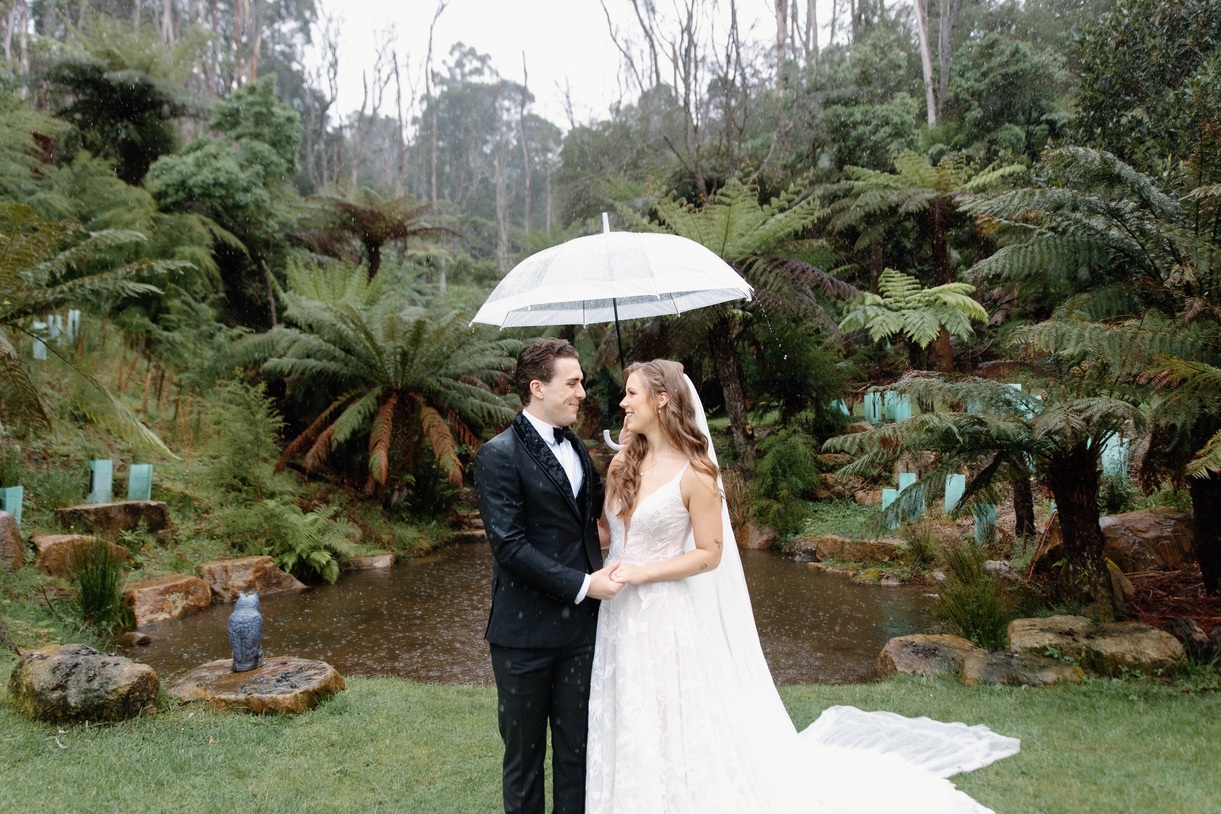 A bride and groom standing together under an umbrella in a lush garden with ferns and a pond, during a light rain.