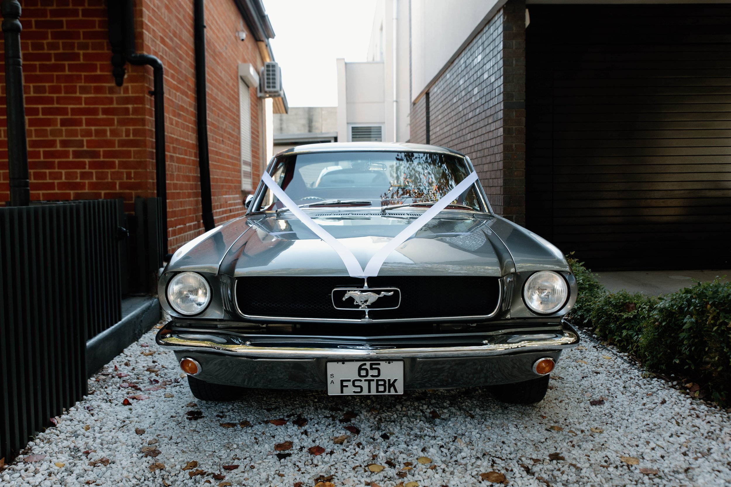 A classic black Ford Mustang parked between brick and white buildings with white ribbons crossing on the hood, and a visible license plate reading '65 FSTBK'.