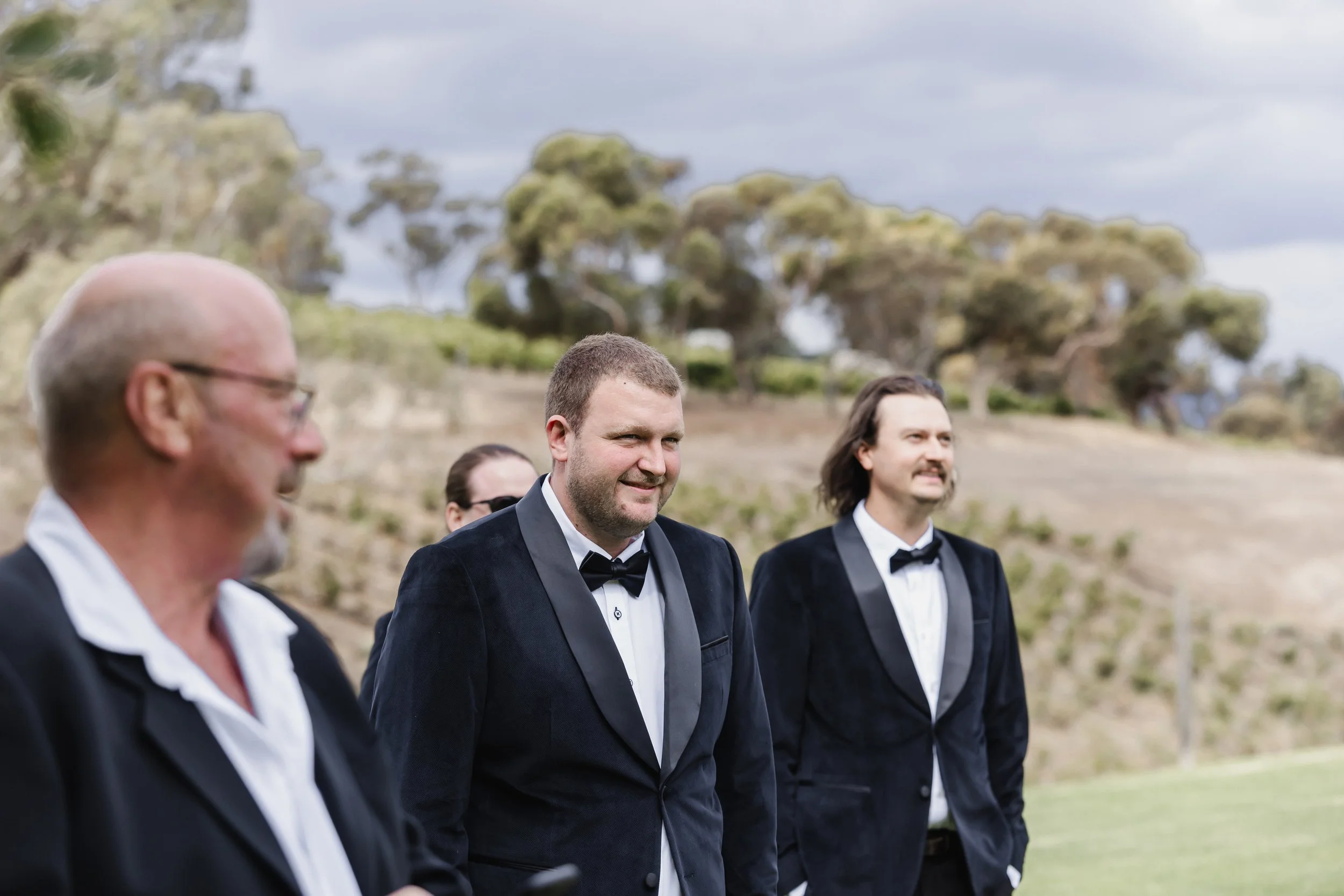 Group of men in tuxedos standing outdoors on a grassy area, with trees and cloudy sky in the background.
