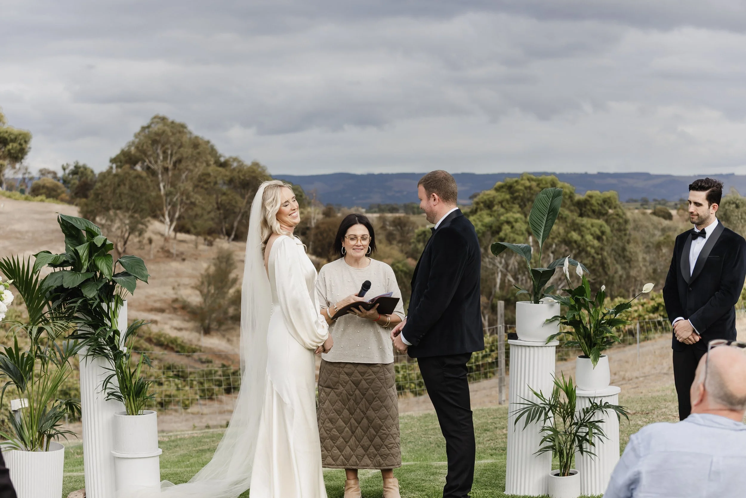A wedding ceremony outdoors with bride and groom holding hands, officiant reading from a book, and guests watching, surrounded by potted plants against a scenic landscape with trees and hills.