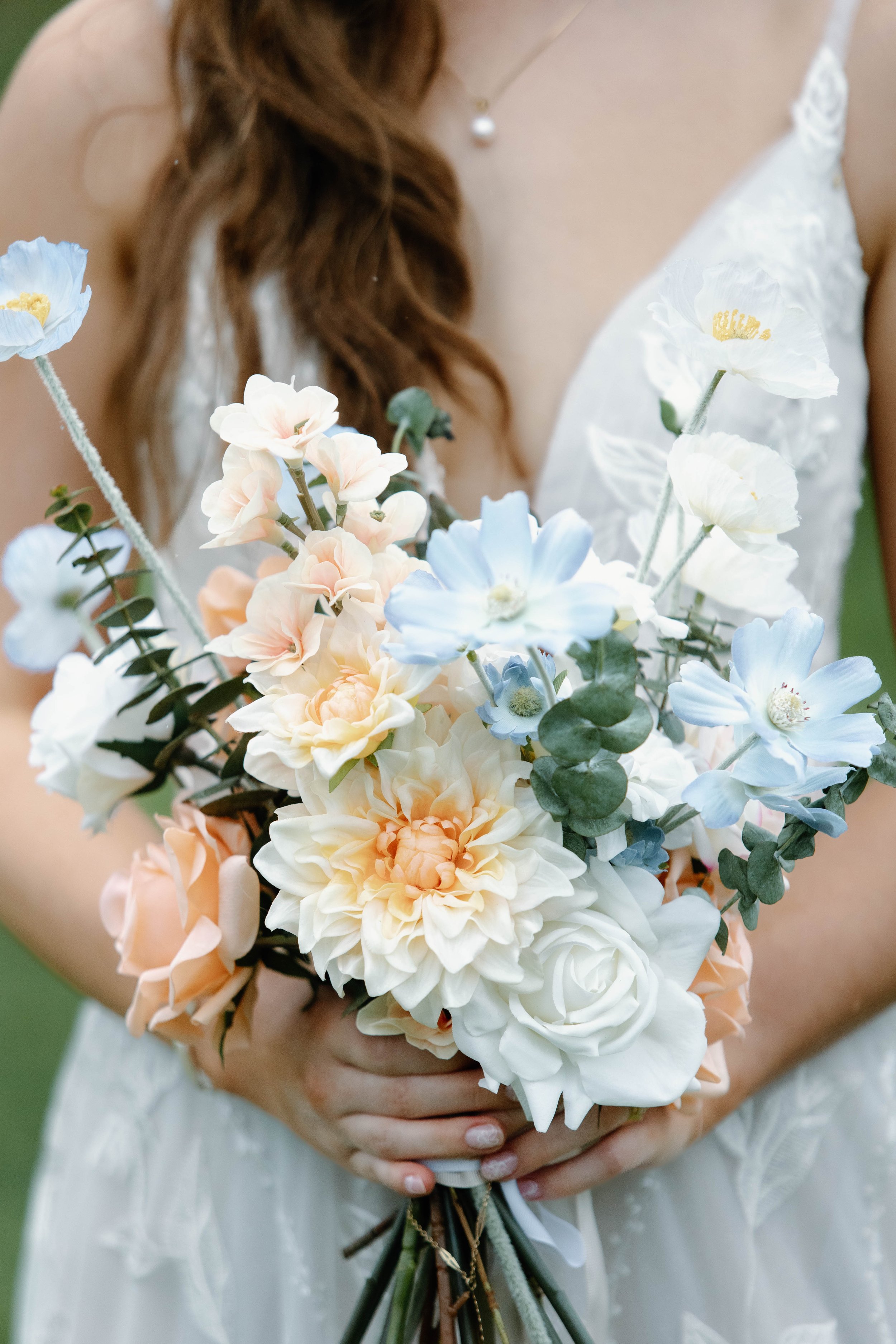 A bride holding a bouquet of pastel-colored flowers, including roses, hydrangeas, and dahlias, with a white lace wedding dress and a pearl necklace.