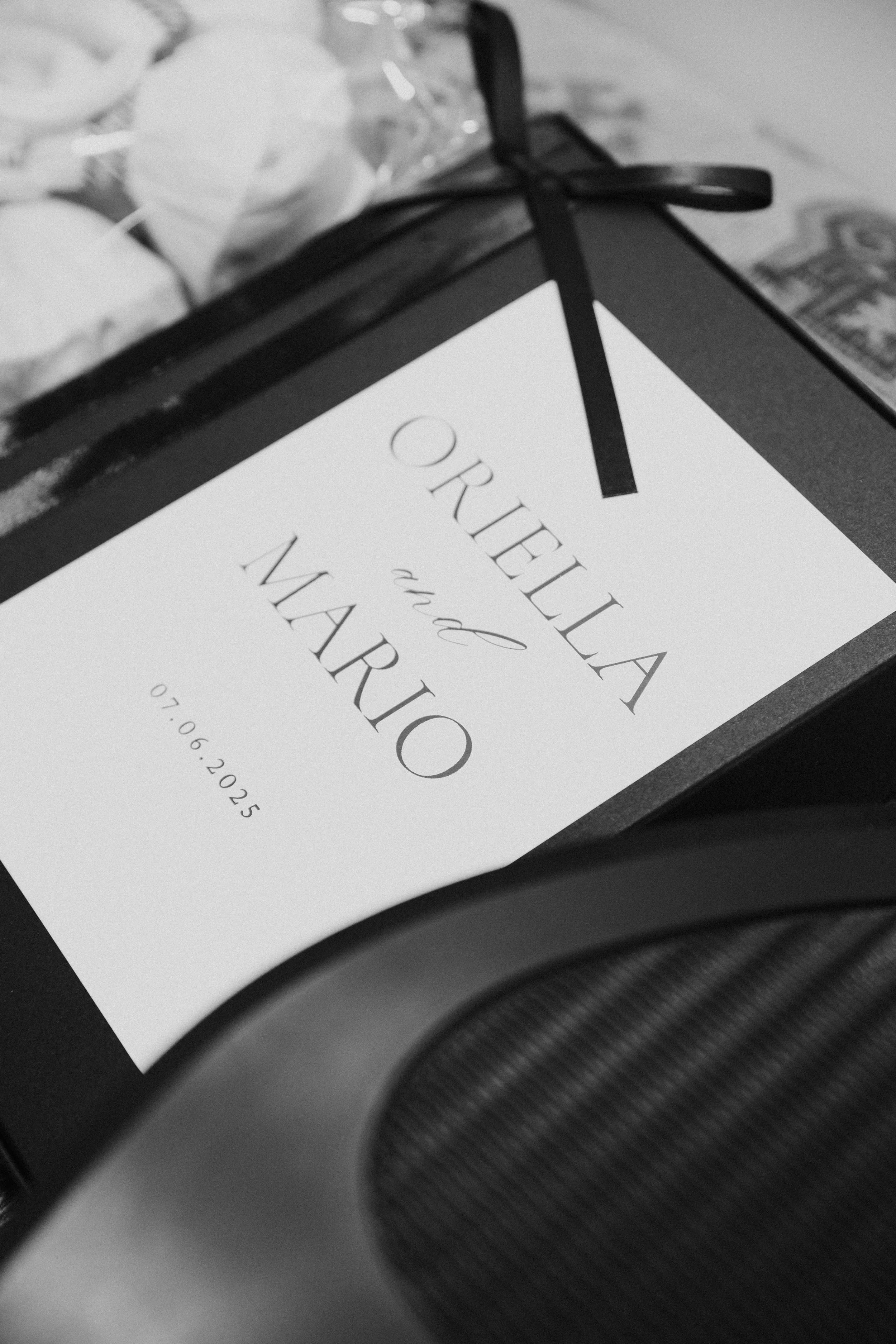 A wedding invitation with the names Orella and Mauro, and the date 07/06/2025, placed on a table with some tissue papers in the background.