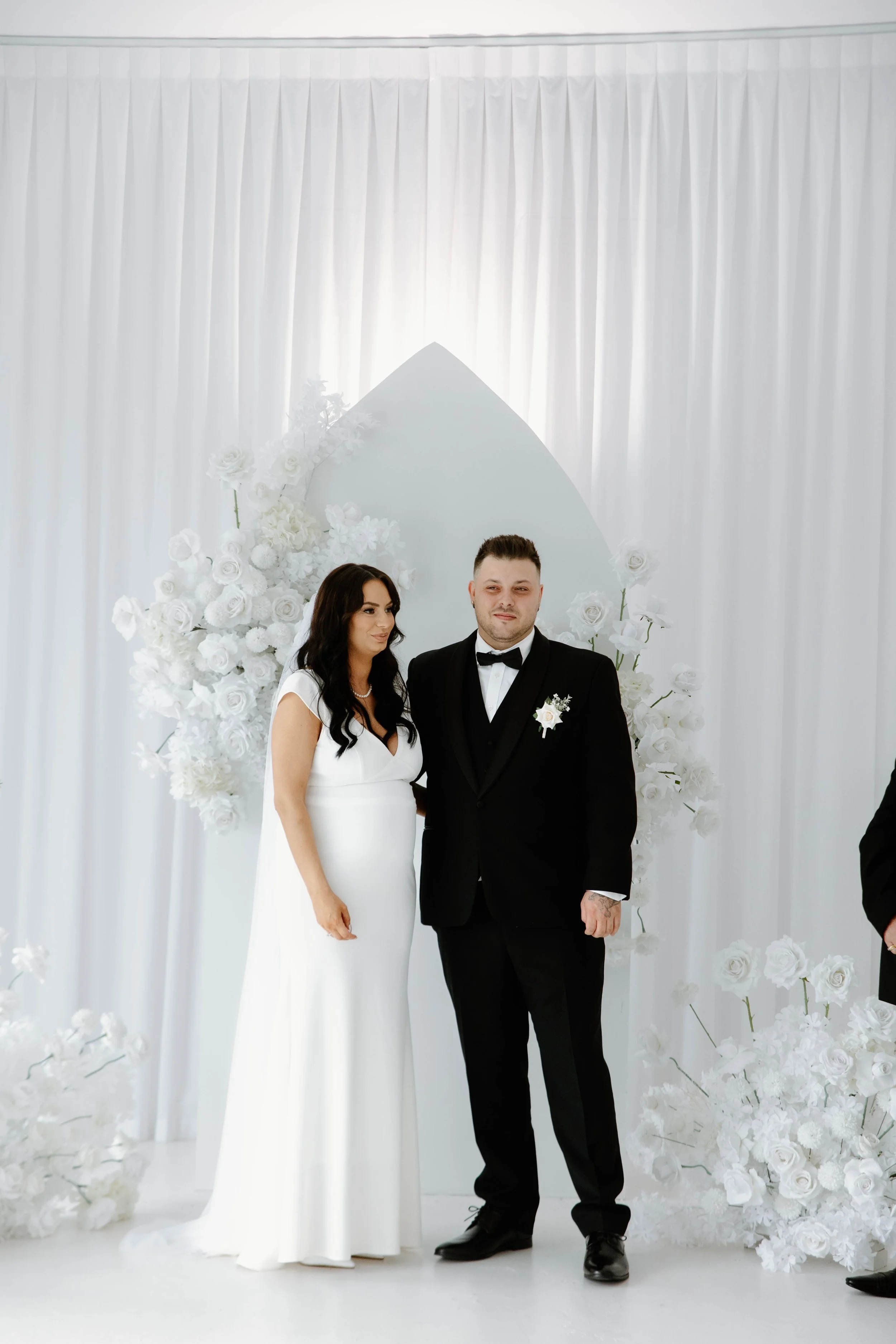 A bride in a white wedding dress and a groom in a black tuxedo standing together during a wedding ceremony, with white floral decorations and drapery in the background.
