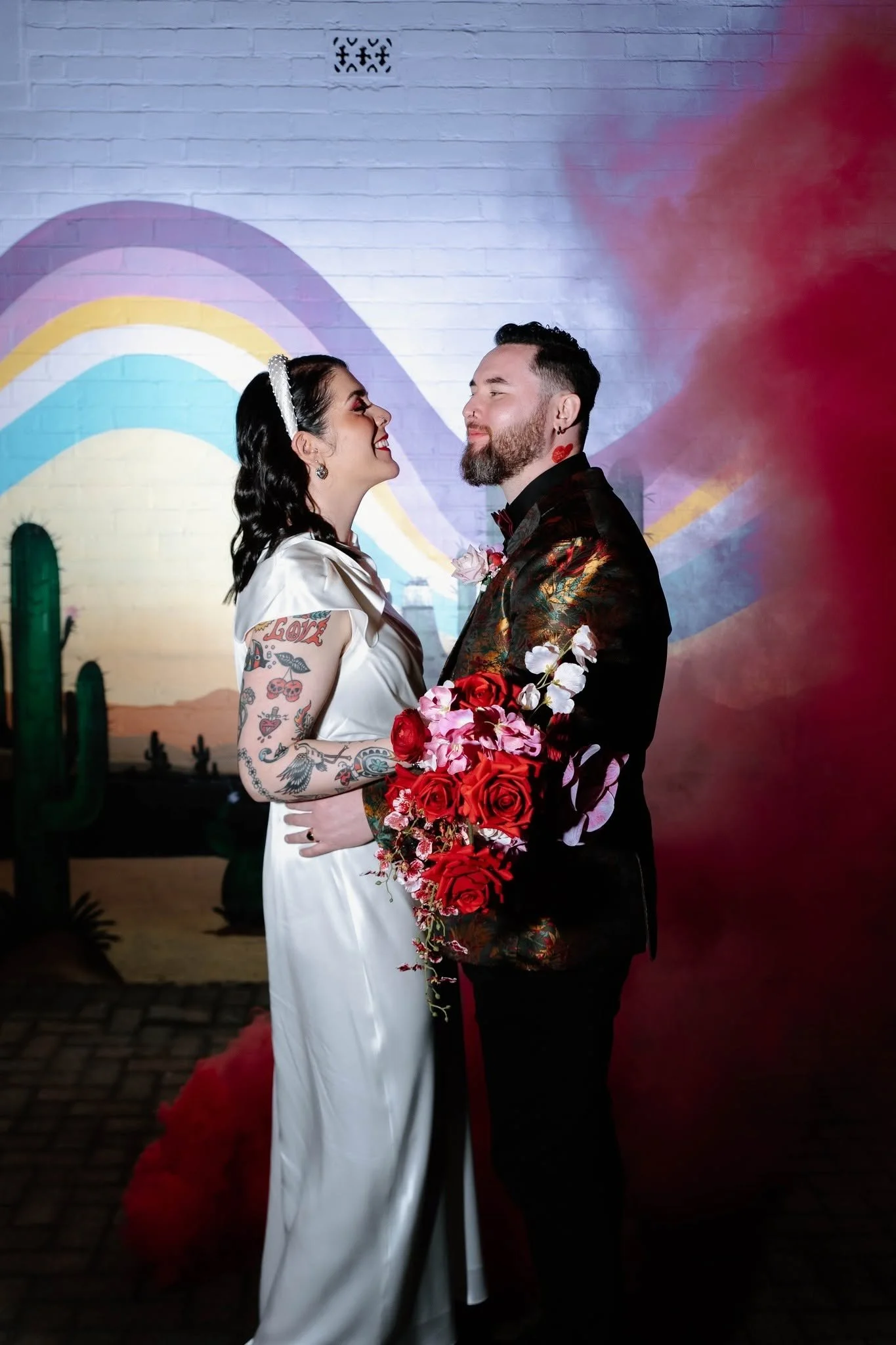 A couple in wedding attire standing face to face, smiling, with a colorful rainbow mural on the wall behind them, and holding a bouquet of red and pink flowers.