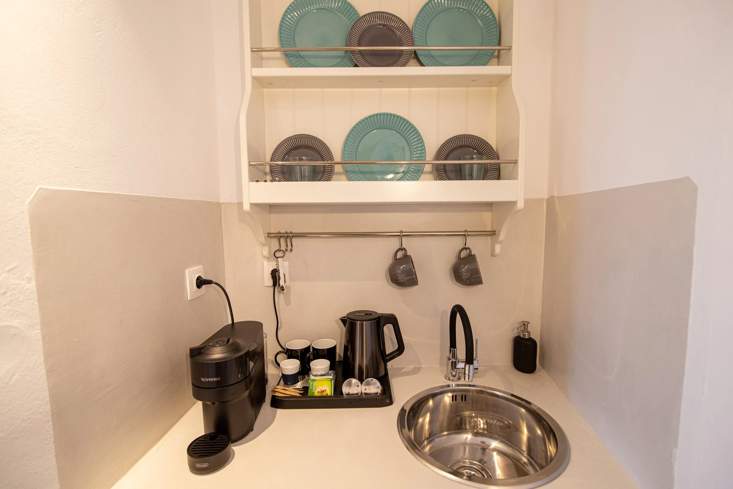 Small kitchenette with a sink, electric kettle, coffee machine, cups, and a shelf holding plates and bowls.