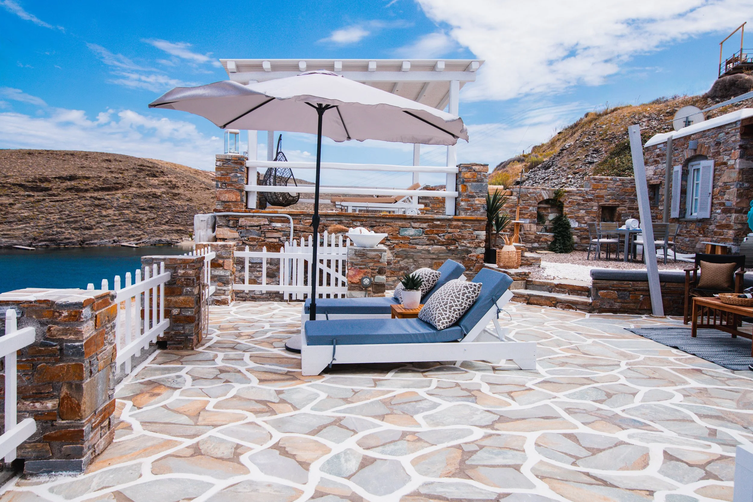 A seaside patio with stone flooring, cushioned lounge chairs, a large white umbrella, and a white fence, overlooking a body of water and rocky hills under a partly cloudy sky.