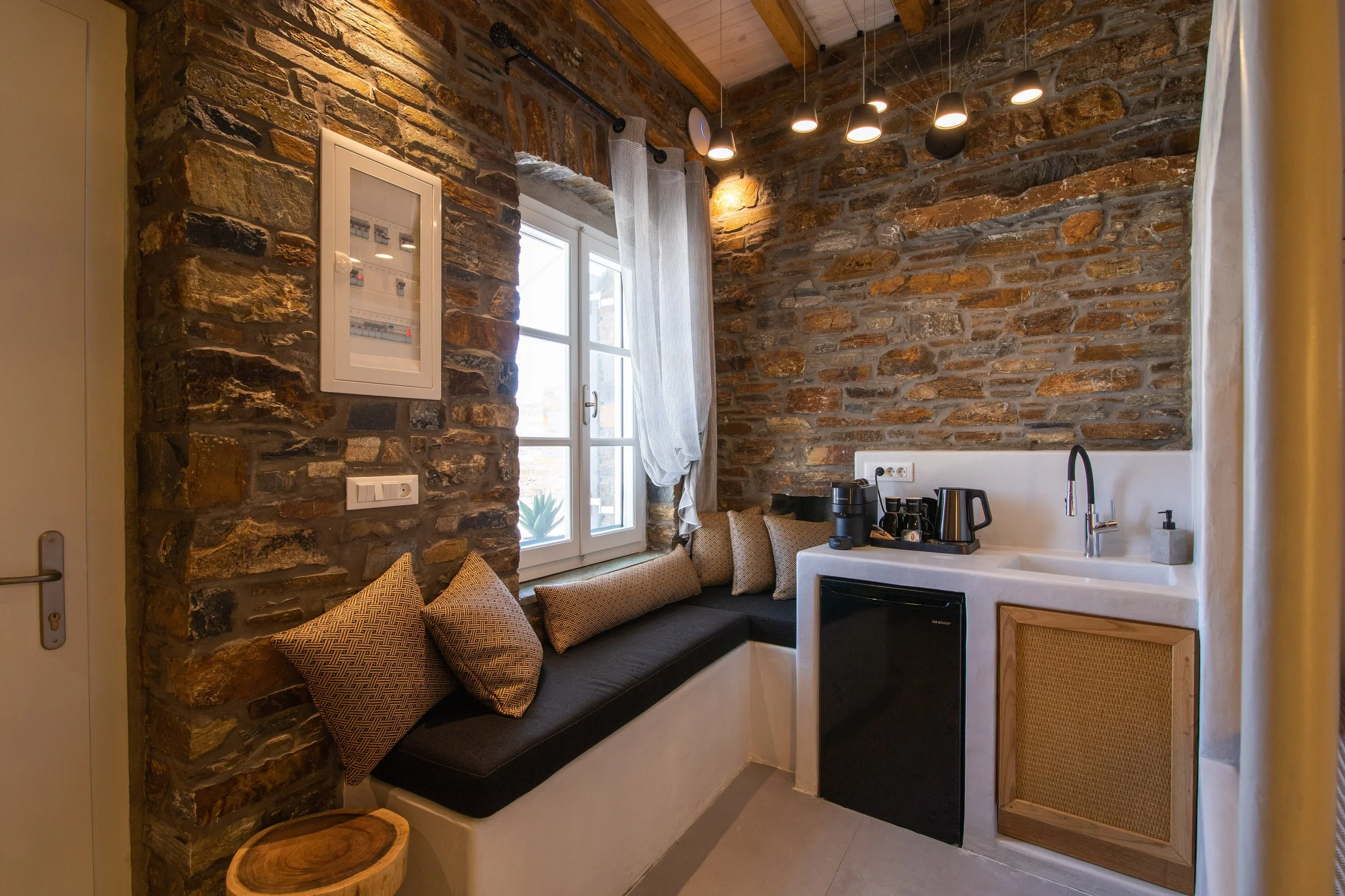 Small kitchen corner with a built-in seating area with beige pillows, brick wall, window with curtain, small refrigerator, coffee maker, kettle, and sink.
