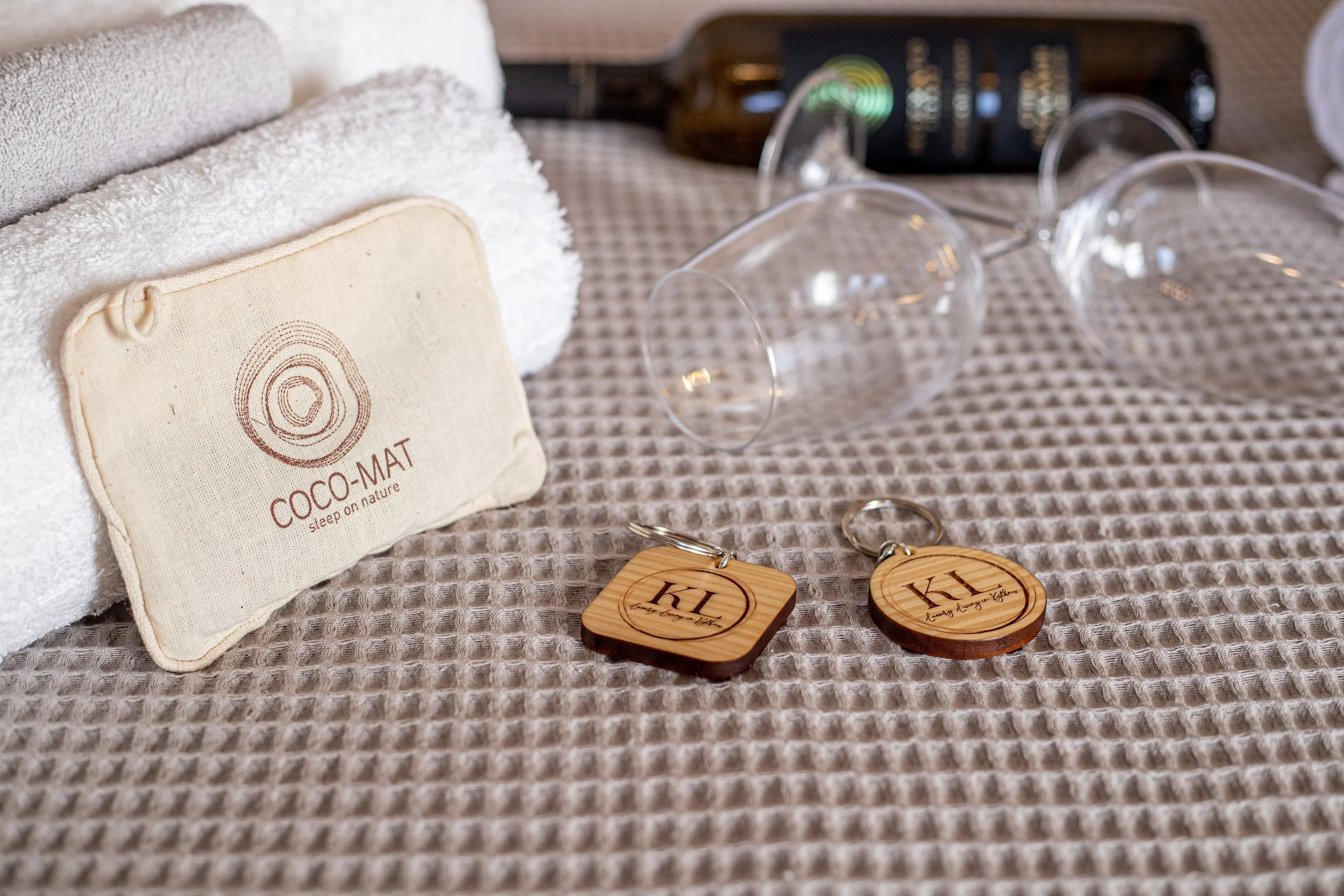 Two wooden keychains with initials KL on a beige textured blanket, alongside two clear wine glasses, a rolled white towel, and a beige pouch labeled 'COCO-MAT sleep on nature'.