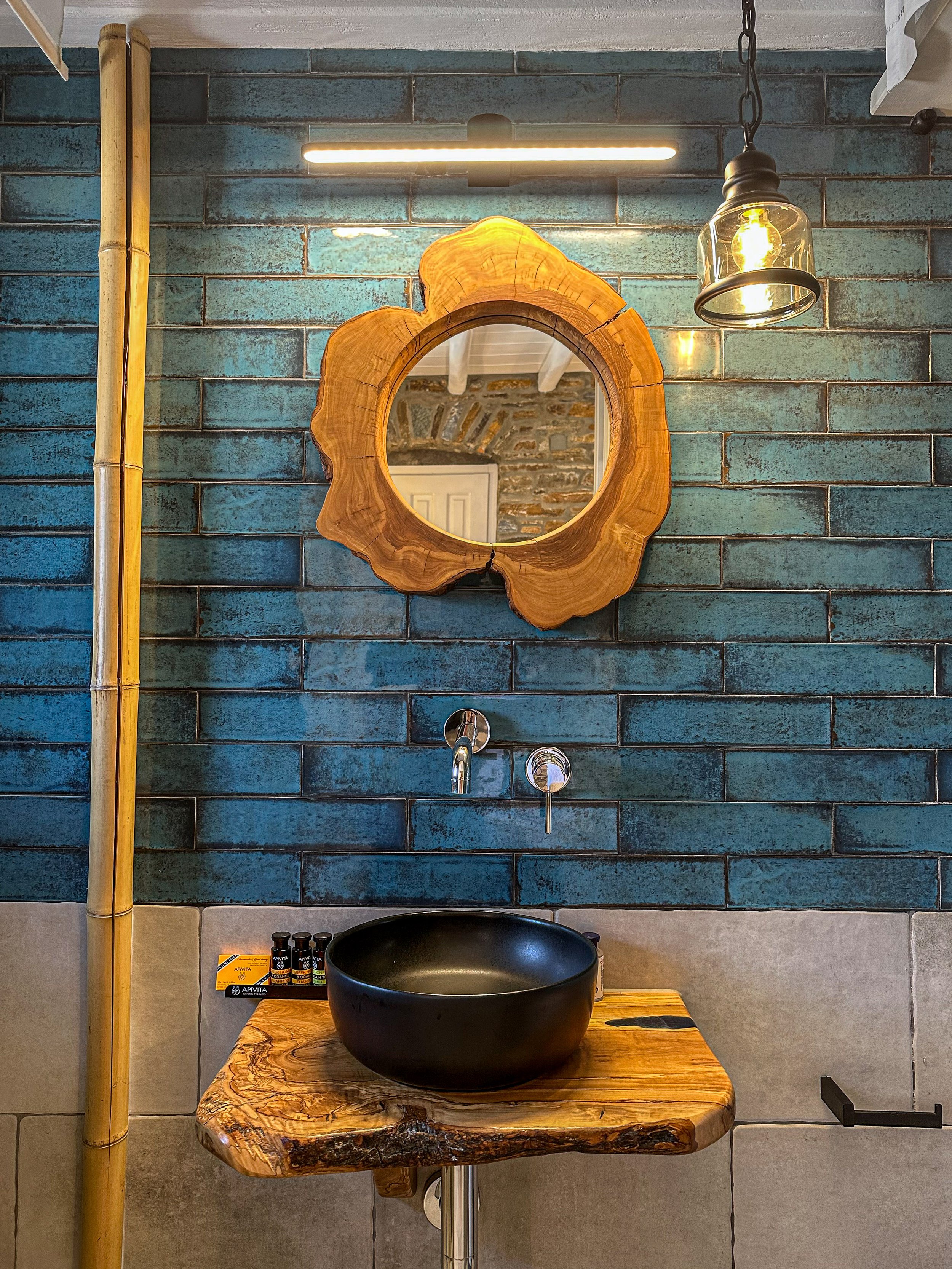 Rustic bathroom with a black vessel sink on a live edge wood countertop, a decorative wooden mirror, and a blue-tiled wall with exposed brick visible through the mirror.