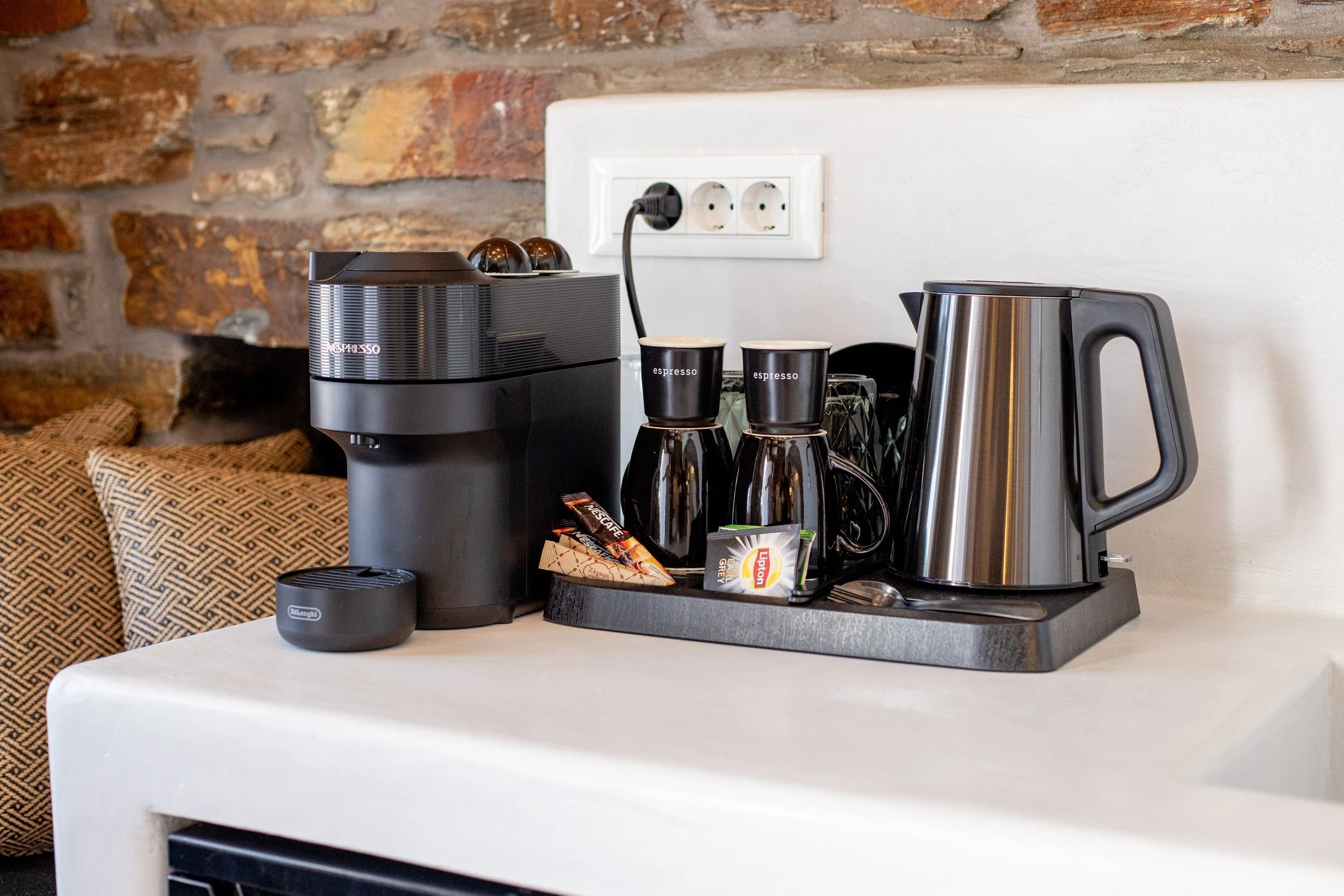 Coffee station with Nespresso machine, two espresso cups, electric kettle, and tea bags on a tray, against a wall with electrical outlets.