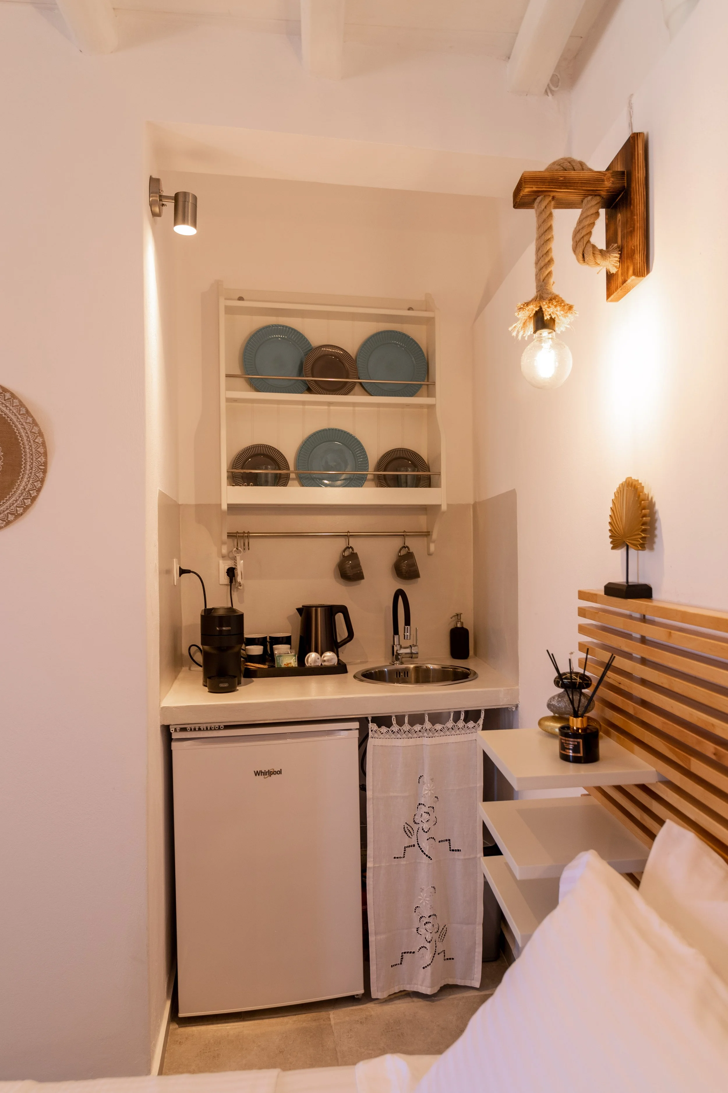 Small kitchenette with a mini fridge, kettle, coffee machine, and hanging mugs, above which are plates on a shelf. A bed with a wooden headboard and decorative pillows is visible in the foreground.