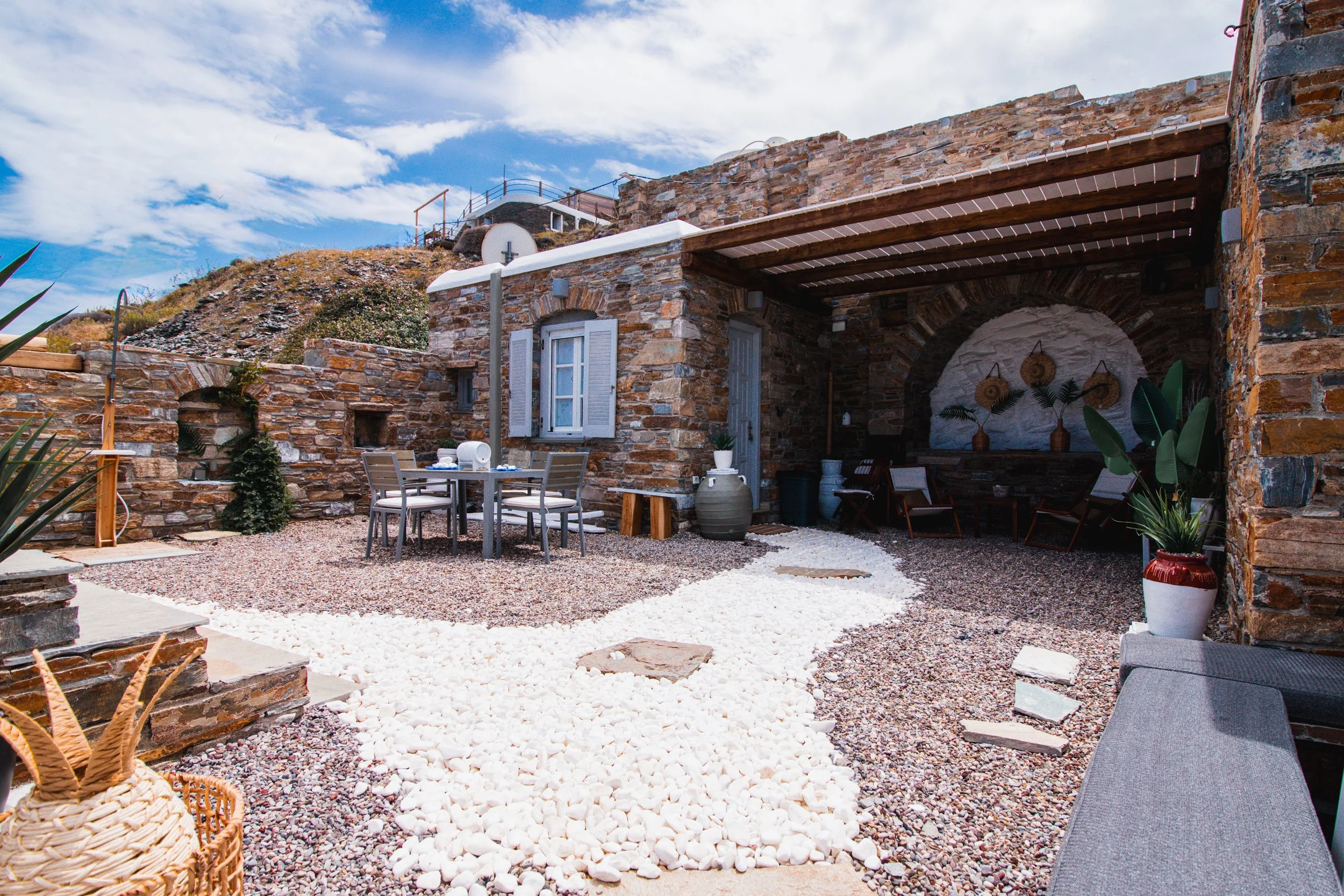 Outdoor patio with brick walls, white gravel pathway, patio furniture, potted plants, and a pergola under a partly cloudy sky.
