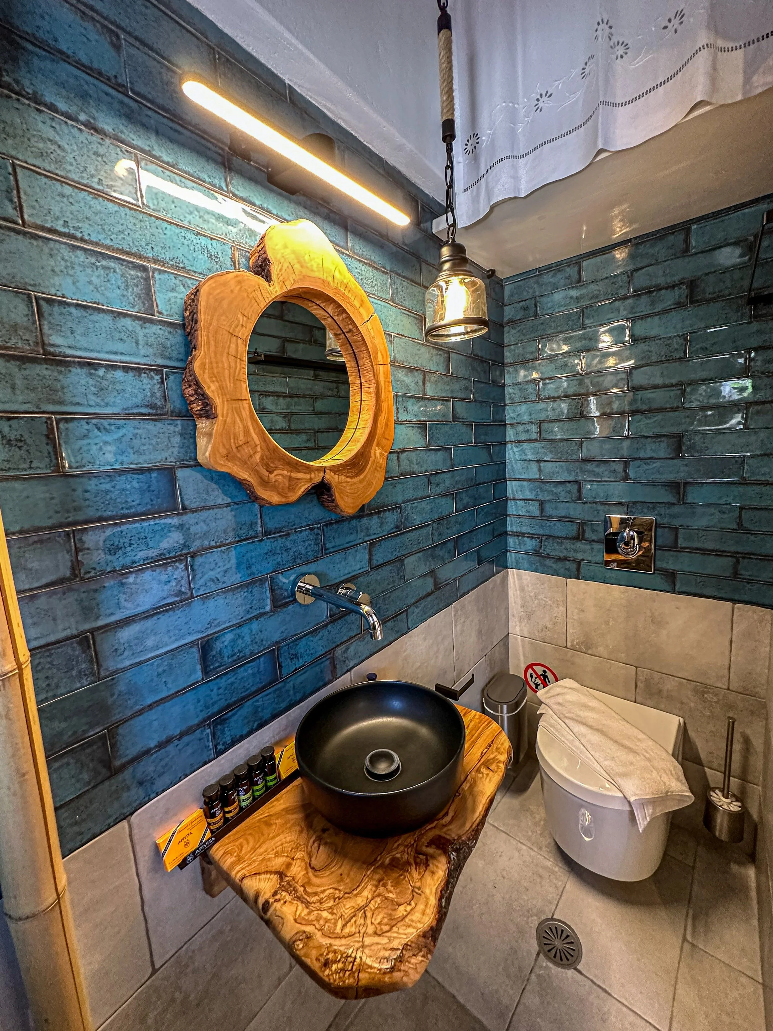 A bathroom with dark teal tiled walls, a round wooden-framed mirror, a wooden countertop with a black vessel sink, bottles of soap or lotion, and a modern toilet with a towel on top. There is a hanging light fixture and a small cylindrical trash can.