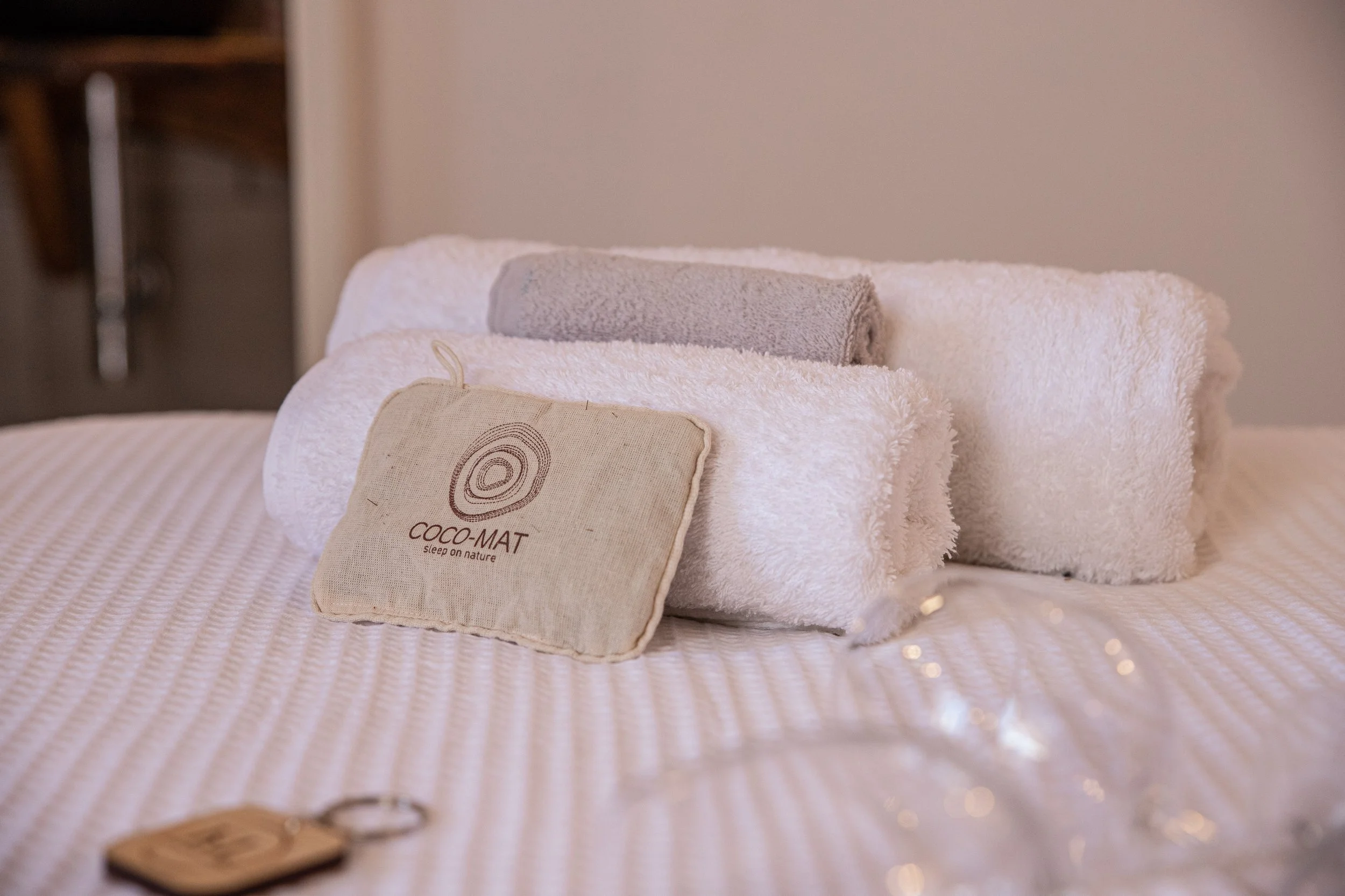 A neatly arranged bed with rolled white towels, a small beige pillow with the logo 'COCO-MAT' and the phrase 'sleep on nature,' and a gray towel on top. A small wooden tray with a bottle and glasses is visible in the foreground.