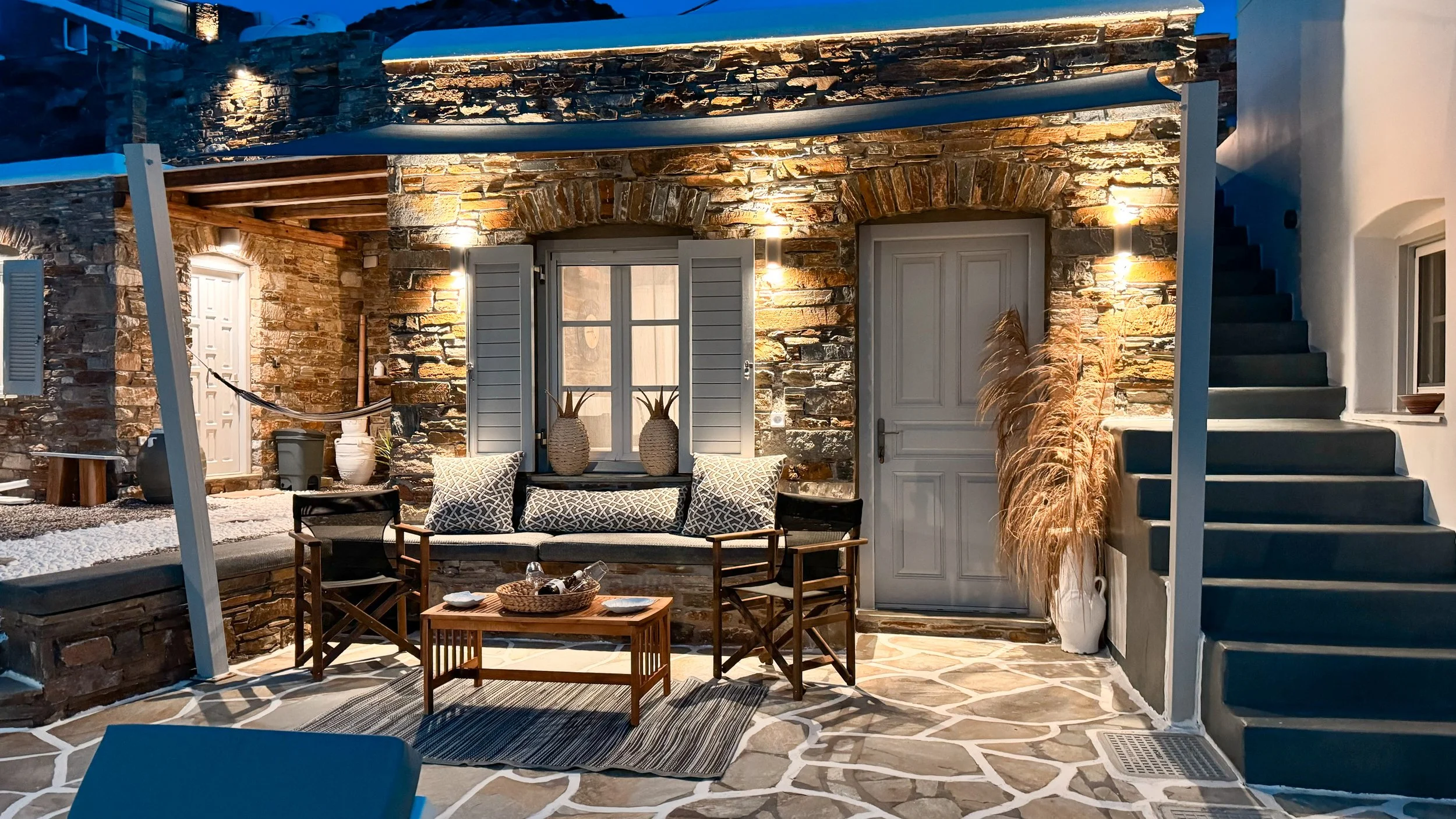 Outdoor patio with stone walls, a bench with cushions, a small wooden table, potted plants, a white door, and stairs leading up on the right side, illuminated by wall-mounted lights at dusk.